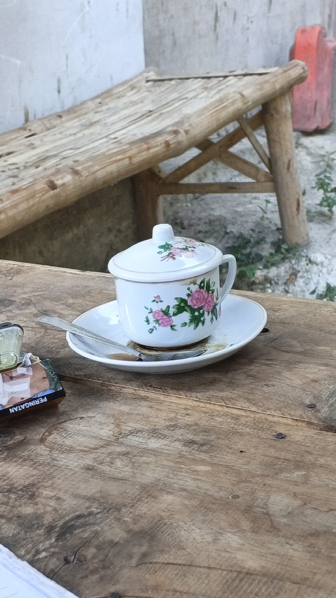 In Rengel, Indonesia, a close-up shot focuses on a floral-patterned teacup with a lid, resting on a saucer atop a rustic wooden table. A small spoon is partially submerged in the dark liquid within the cup. To the left of the cup, a cigarette pack with Indonesian text "PERINGATAN" (Warning) is visible, along with a small, clear lighter. In the background, a simple wooden bench made of rough-hewn logs and branches sits near a textured concrete wall. The scene evokes a sense of quiet contemplation or a pause from daily activities.