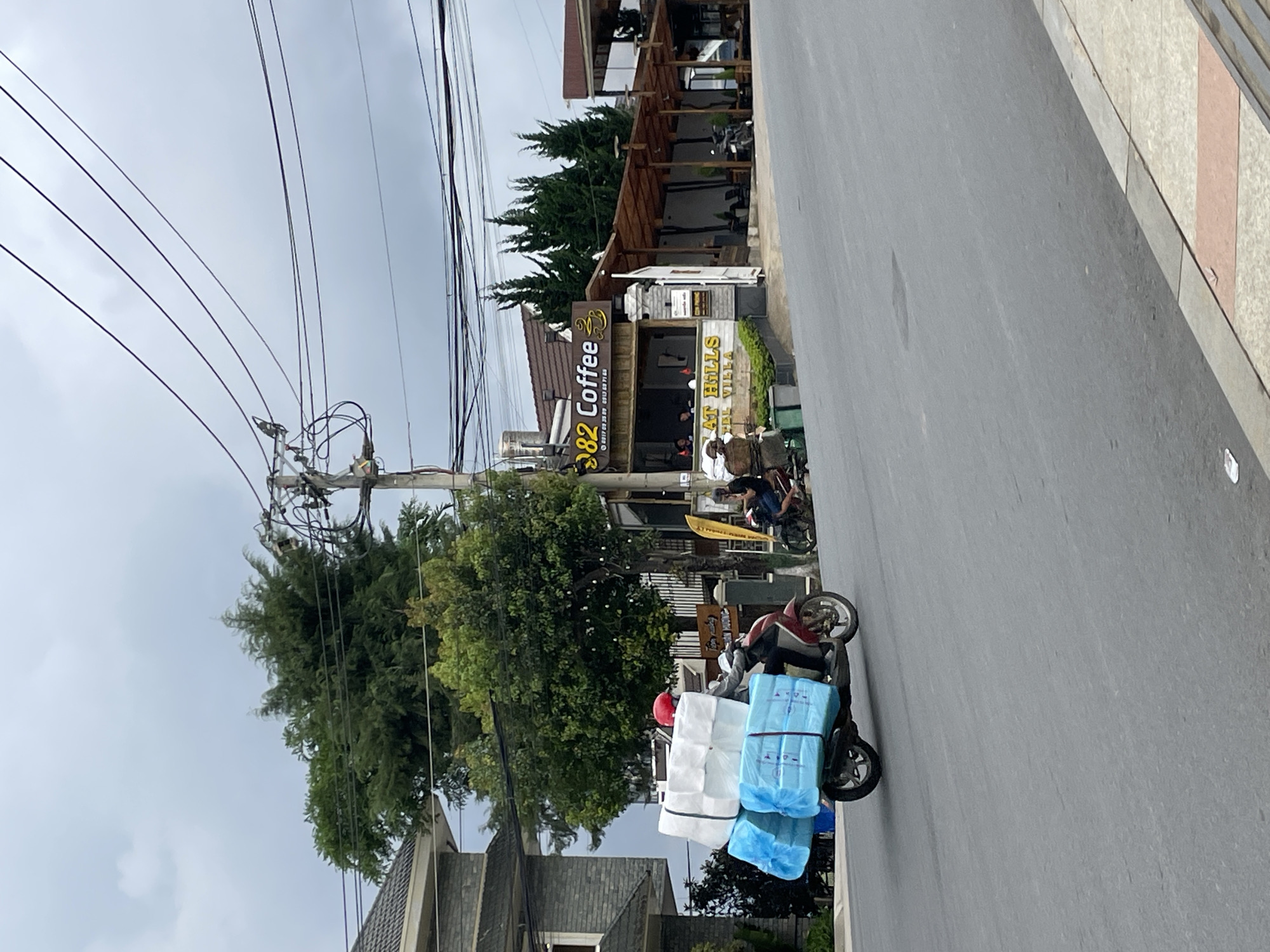 In Lâm Viên - Đà Lạt, Vietnam, a person wearing a red helmet rides a motorcycle laden with a significant cargo. The motorcycle is carrying stacks of blue and white packaged goods, secured on the rear rack. The rider, dressed in a dark jacket and pants, is positioned behind the handlebars, navigating the paved road. The environment is characterized by a cloudy sky, overhead power lines, and trees bordering the road. To the right of the road, a coffee shop with an outdoor seating area and a sign that reads "82 Coffee" is visible. Other motorcycles are parked near the coffee shop, and two individuals are seated at a table. A residential building with a gray tiled roof is present on the left side of the image.