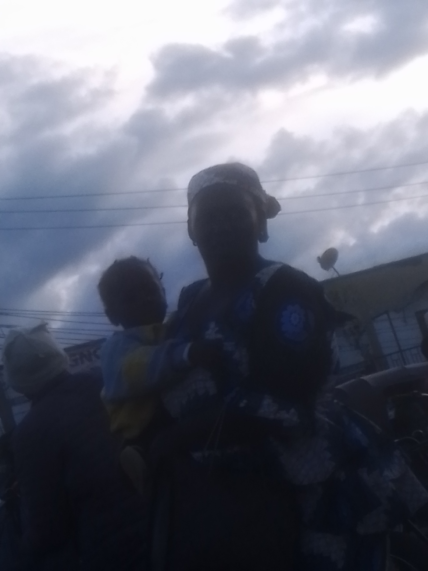 The image shows a dark, blurry scene, likely taken outdoors in Gwong, Nigeria, with a cloudy, overcast sky. In the foreground, a woman is holding a child. The woman is wearing a headscarf and a patterned dress. The child is wearing a blue and yellow outfit. To the left of the woman and child, another person is visible wearing a light-colored head covering. In the background, there are buildings and what appear to be power lines. The overall lighting is dim, suggesting it may be late afternoon or early evening, or a very cloudy day. The image is of poor quality, with significant blur and low resolution.