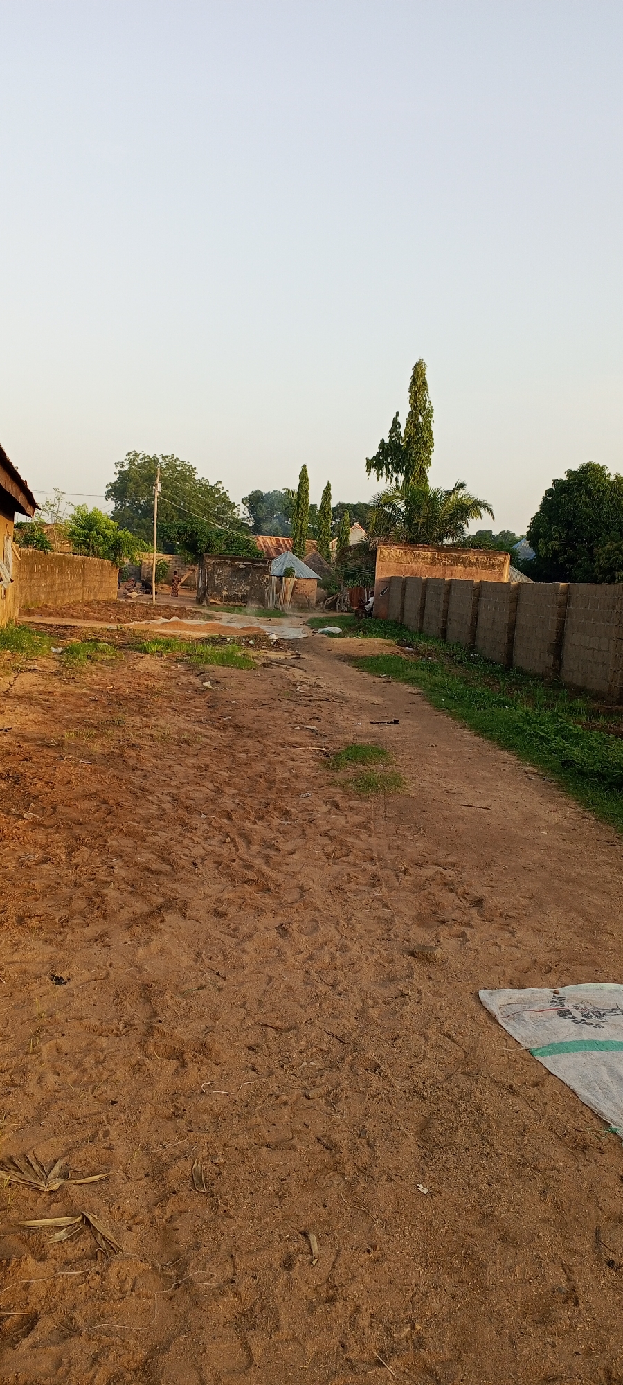 A dirt road passes through a village in Jalingo, Nigeria. The road is unpaved and covered in sand and footprints. On either side of the road are houses and walls made of concrete blocks. Some of the houses have thatched roofs, while others have corrugated metal roofs. There are trees and vegetation scattered throughout the village. In the foreground, a white tarp with green trim lies on the ground. The sky is clear and pale blue, suggesting it is daytime.