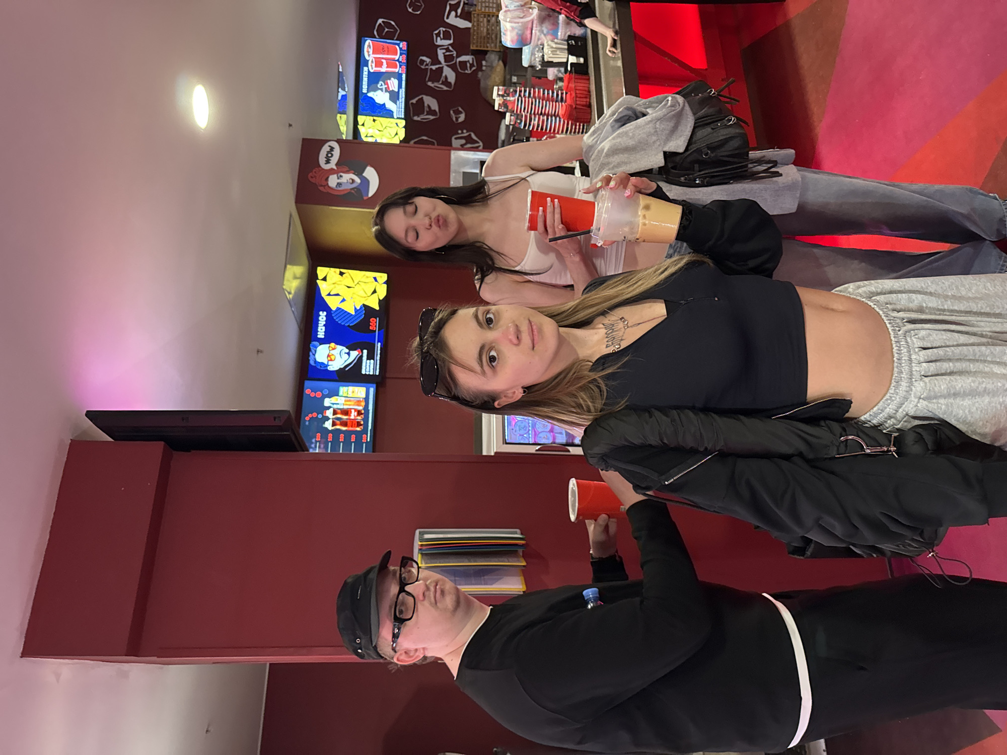 The image depicts a group of three young adults inside a brightly lit, modern establishment with red walls and a vibrant pink and red patterned floor, suggestive of a cinema lobby or entertainment venue, possibly located in Moscow, Russia. In the foreground, a woman with long blonde hair wears a black zip-up jacket over a black cropped top and grey sweatpants, holding a plastic cup with a light brown beverage. To her right, a woman with dark hair and a white tank top is in the middle ground, holding a red plastic cup. To the left, a man wearing a black baseball cap, glasses, and a black sweatshirt holds a red plastic cup. Behind them, digital screens display colorful graphics and text, and a counter area with drinks and snacks is visible. The overall atmosphere is casual and social.