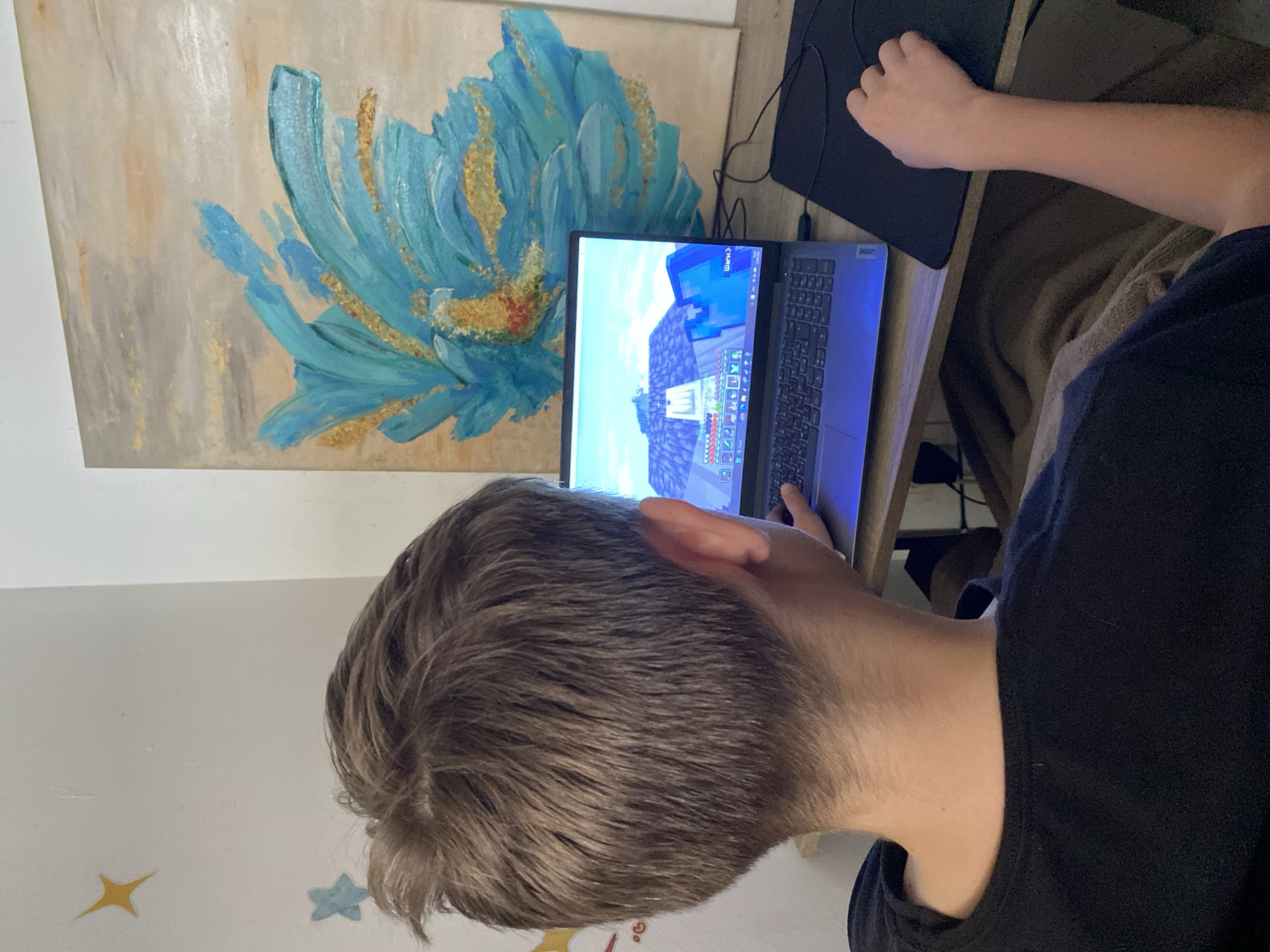 A young person is seated at a desk, focused on a laptop displaying the game Minecraft. A painting with a large blue and gold flower design is visible behind the laptop. A black mousepad sits to the right of the laptop. The young person is wearing a black shirt. The wall behind the person has small star decorations. The location of the city of Trebesin, Montenegro is unknown, therefore, it cannot be determined whether this image comes from Trebesin, Montenegro.