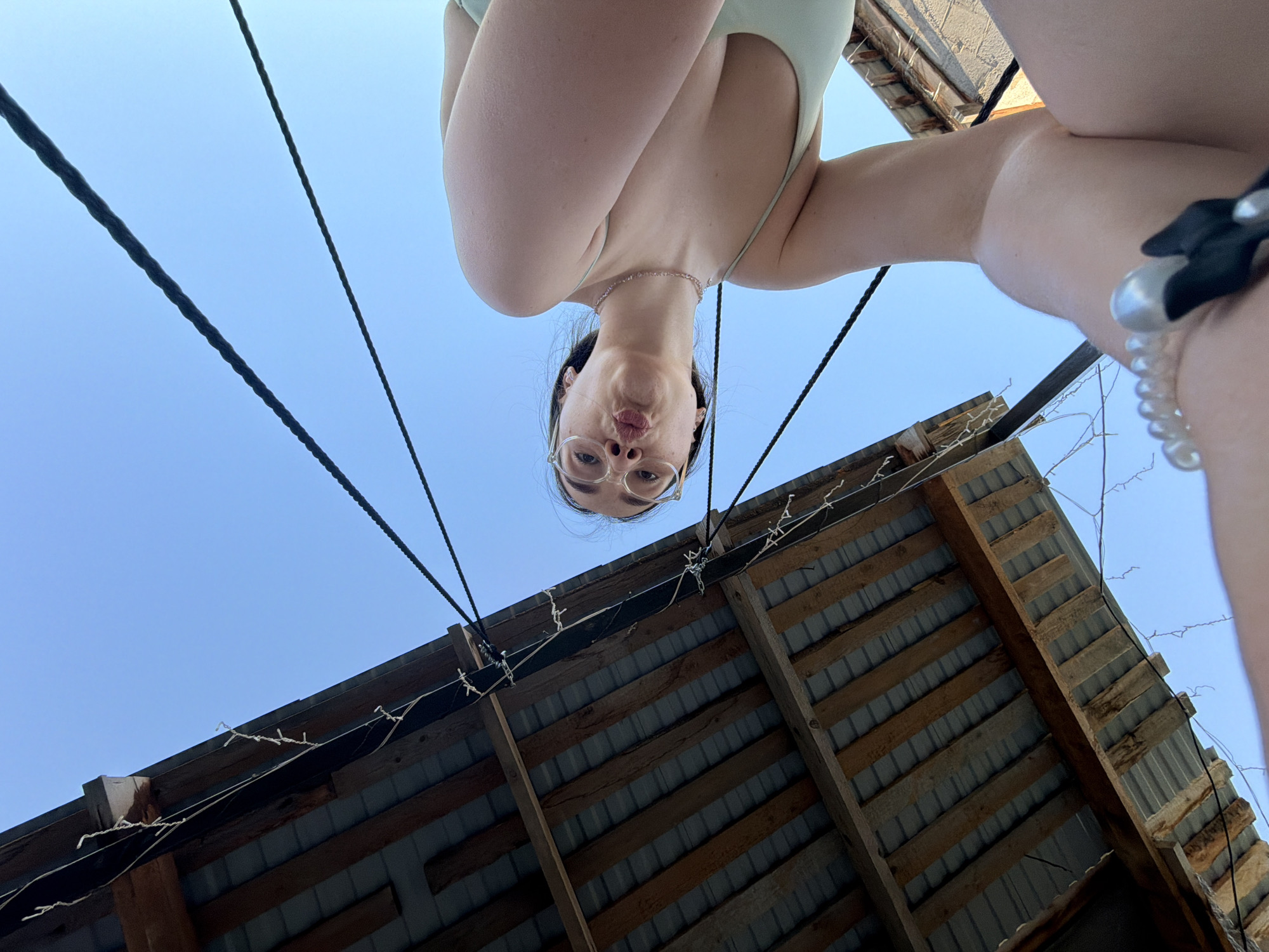 The image shows a person viewed from below, standing above the camera. The person has fair skin, wears glasses, and makes a kissing face. They are wearing a light-colored top and a necklace. In the background is a roof made of wood and corrugated metal, along with dark cables strung across the blue sky. White decorative lights are draped along the edges of the roof. The location is unknown; there is no visible evidence indicating it's Chaikovsky, Russia.