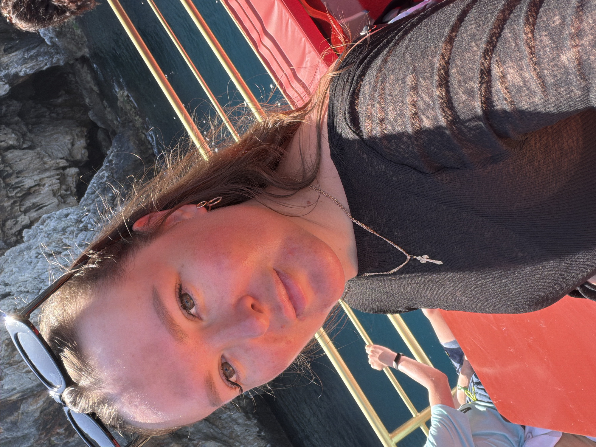 A young woman is the central subject of this brightly lit outdoor portrait, taken on what appears to be a boat. She has long brown hair, brown eyes, and a fair complexion, showing some natural redness, possibly from sun exposure. She wears a black, semi-sheer long-sleeved top and a silver chain necklace with a cross pendant. Black sunglasses are perched on her head. She looks directly at the camera with a neutral expression.
The setting is under clear, strong sunlight, casting distinct shadows on her shirt and creating warm highlights on her hair and face. The boat's features include a prominent red deck and golden railings, visible both behind and in front of her. Beyond the boat, a calm body of deep blue-green water is visible, stretching alongside rugged, rocky cliffs or coastline, consistent with a scenic coastal area like Alanya, Turkey. In the lower right corner, an arm of another individual, possibly a child, wearing a black wristband, can be seen holding onto a golden railing. The scene depicts a sunny day out on the water, likely during an excursion. There is no visible text in the image.