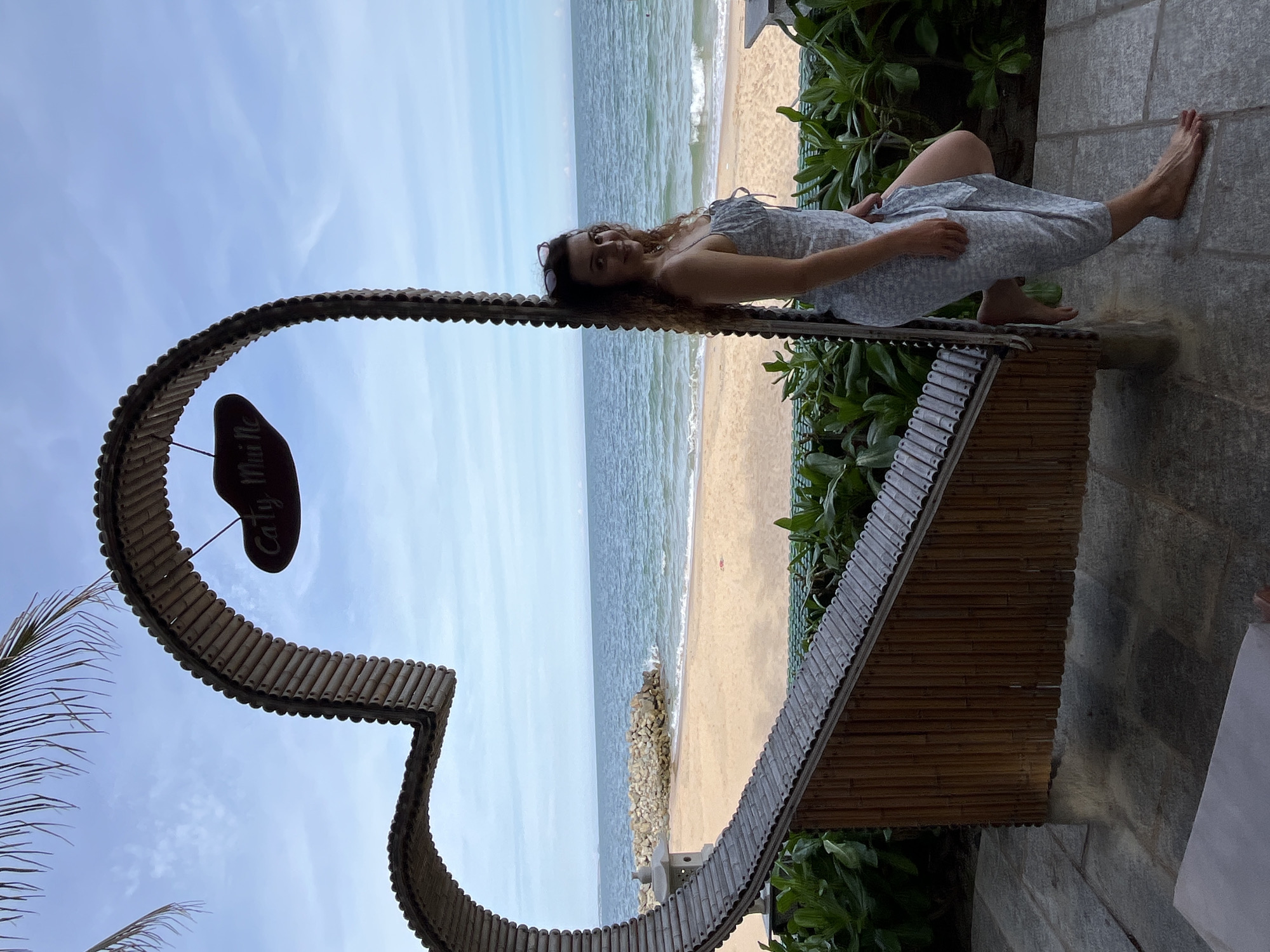 A young woman is standing next to a large heart-shaped bamboo structure at a beach in Mũi Né, Vietnam. The heart structure frames a view of the ocean, a sandy beach, and some rocks. The woman is wearing a light-colored floral dress and sunglasses are perched on her head. She is leaning against the bamboo structure with one leg bent. The sky is partly cloudy and the sea is blue. Lush green plants are visible in the foreground. A sign hangs from the top of the heart structure, which appears to read "Caty Muine."