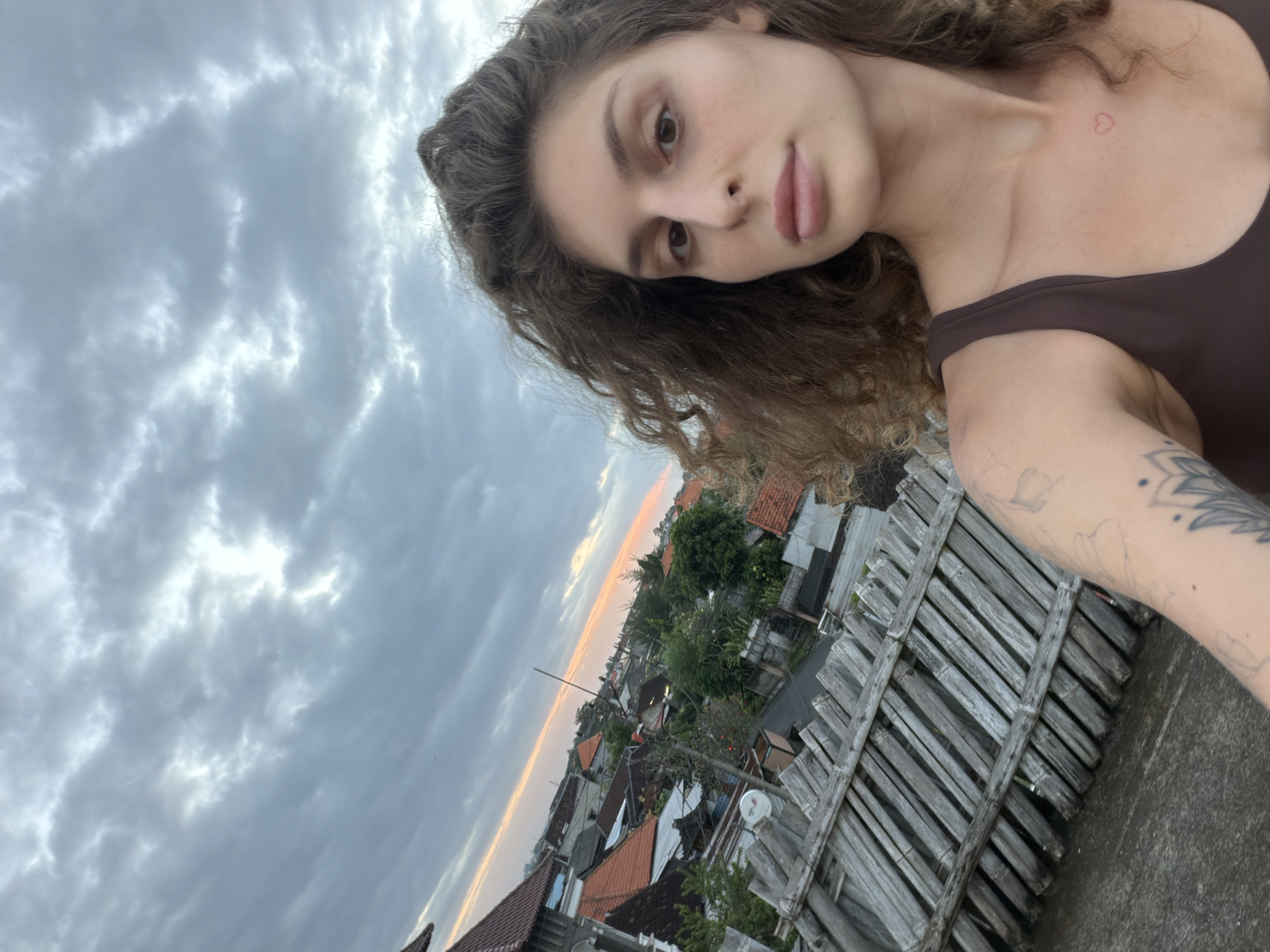 A selfie image shows a woman with brown curly hair, a heart tattoo on her upper chest, and a brown tank top. She has fair skin and her face is turned toward the camera. The background reveals a cityscape with a variety of structures, including buildings with orange tile roofs, amidst lush greenery. A weathered wooden fence is also visible. The sky above is overcast with shades of gray. This image was taken in North Kuta, Indonesia.