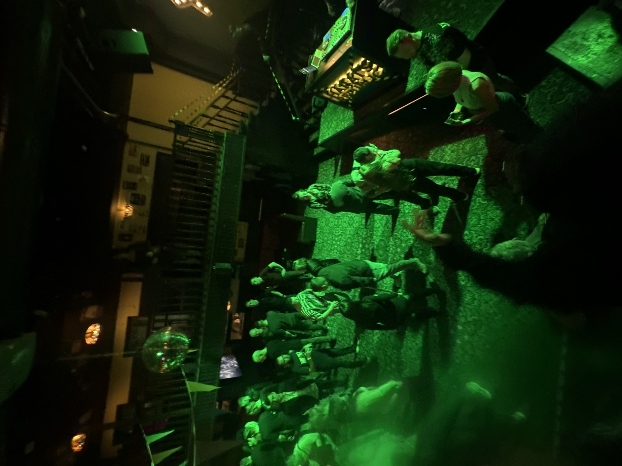 The image depicts what appears to be a bar or club setting with a crowd of people. The location is possibly in Saratov, Russia. There is a staircase visible in the background, along with a disco ball. Individuals are standing and interacting in the space. A bar is present in the upper portion of the image. The lighting is predominantly green.