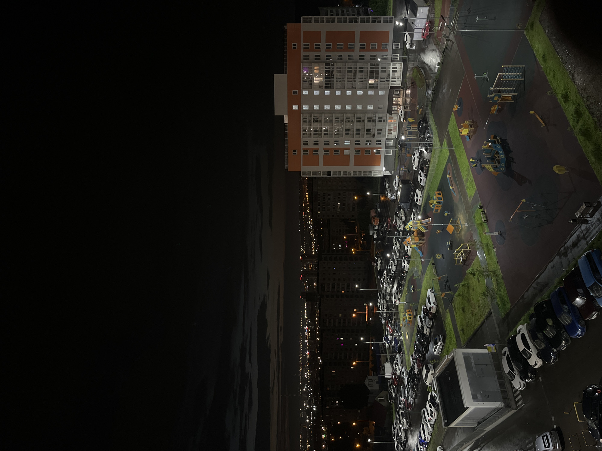 This is a nighttime aerial photograph of a residential area in Krasnoyarsk, Russia. The scene features a modern apartment building with orange and white accents and numerous illuminated windows. Adjacent to the building is a playground with various structures, including slides and climbing frames, situated on a dark, multi-colored surface with patches of grass. A large parking lot is filled with numerous cars, many of which are white. The time of day is clearly night, indicated by the dark sky, illuminated windows, and streetlights. The ground appears wet, suggesting it might have rained recently or the weather is damp. In the background, a city skyline with many lights stretches across the horizon, hinting at a densely populated urban environment. The overall mood is quiet and possibly tranquil, illuminated by artificial light against the darkness. No text is visible in the image.