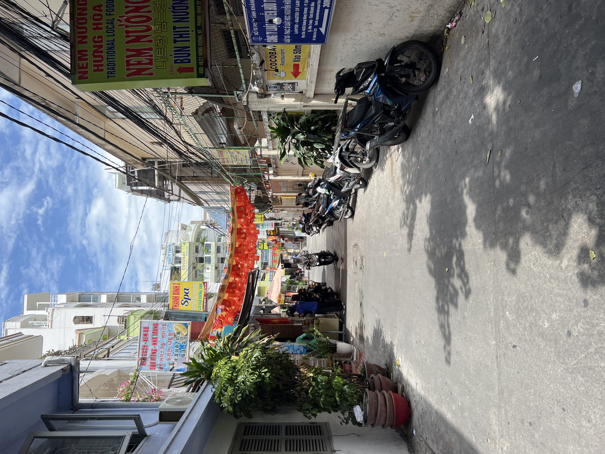 This is a ground-level, eye-level, medium-shot photograph of a narrow, paved street in Nha Trang, Vietnam. The street is lined with buildings of varying heights and architectural styles. The buildings are painted in muted colors such as white, beige, and light blue, with some green accents. Electrical wires are strung across the street overhead.
On the left side of the street, there are potted plants and a white building with louvered shutters. Red paper lanterns hang across the street. Signs in Vietnamese and English advertise services such as "Spa," "Foot & Body Massage," and "Restaurant."
The right side of the street features more signs, including one for "NEM NUONG HUNG HOA TRADITIONAL LOCAL FOOD." This sign has Vietnamese and Korean text. Another sign, also in Vietnamese, advertises a law office. A restaurant called "COCOBA" is also visible.
Several motorcycles are parked along the right side of the street. Two people are standing near a stall on the left side, and one person is riding a motorcycle down the street towards the viewer. The ground is paved with asphalt and is somewhat littered with leaves and small pieces of debris. Shadows of trees are cast across the street. The sky is blue with white clouds.