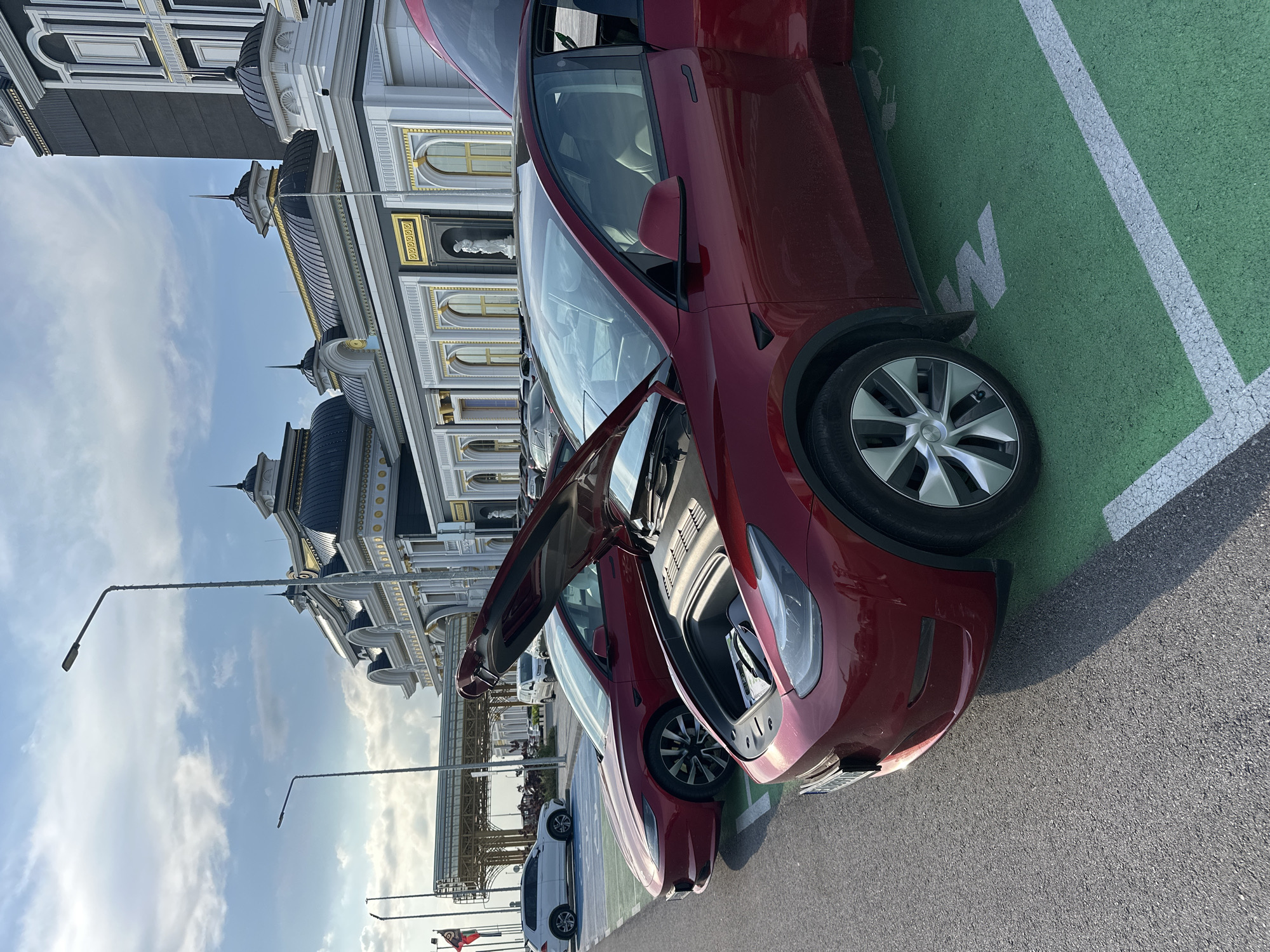 The image depicts an outdoor parking lot in Kapitan Andreevo, Bulgaria, featuring two red Tesla electric vehicles. In the foreground, a red Tesla Model Y is parked in a green-marked electric vehicle charging bay, indicated by a white charging symbol and a partial white 'M' or 'W' on the green pavement. The vehicle's front trunk (frunk) is open, revealing a storage compartment containing a dark, rectangular object resembling a folder or manual. The driver's side door is also open. Directly behind the Model Y, a red Tesla Model 3 is parked with its hood open.
The background features a large, ornate building exhibiting classical architecture, characterized by dark grey/black domed roofs, white facade elements, gold trim, arched windows, and decorative cornices. A white statue is visible in a niche on the building's facade. Further to the left, a white SUV is parked, and a covered walkway structure with metal framework is visible. Several tall streetlights stand throughout the scene. The sky is partially cloudy, and sunlight casts shadows on the asphalt, indicating daytime. No people are visible in the frame.