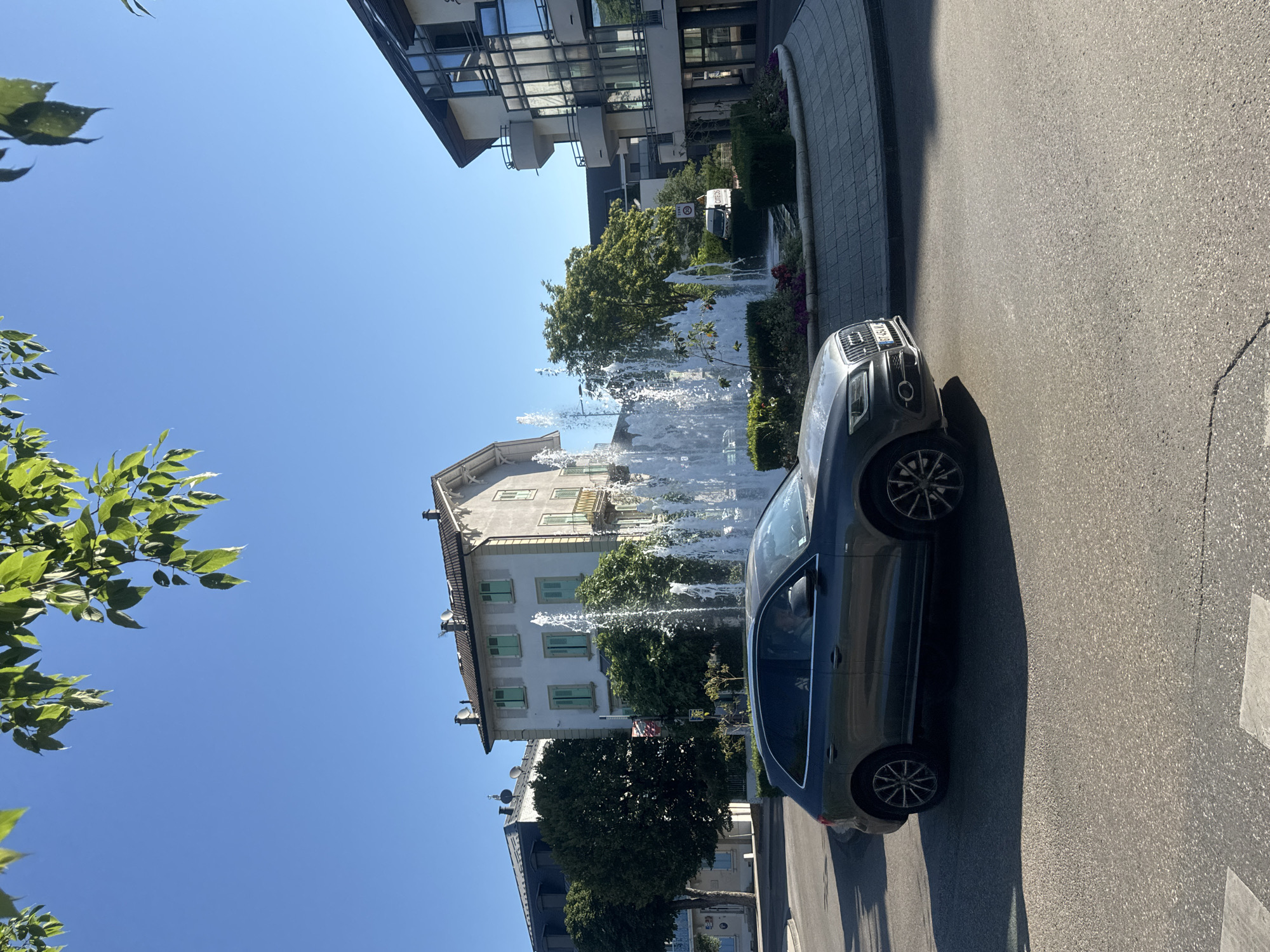 A dark gray Audi SUV is captured in motion on a paved road in Divonne-les-Bains, France. The vehicle is positioned in the foreground, on the right side of the frame, partially obscuring a circular fountain with multiple water jets. Behind the fountain, several buildings are visible. To the left of the fountain stands a light-colored, multi-story building with green shutters and balconies. Further to the left, another building with a dark roof is partially visible, and a cluster of trees is situated between the two buildings. To the right of the fountain, a modern building with a glass facade and balconies is present. The scene is illuminated by bright sunlight, casting shadows on the road. The sky is clear and blue, with the leaves of a tree hanging into the upper portion of the frame. A speed limit sign reading "ZONE 30" is visible near the fountain.