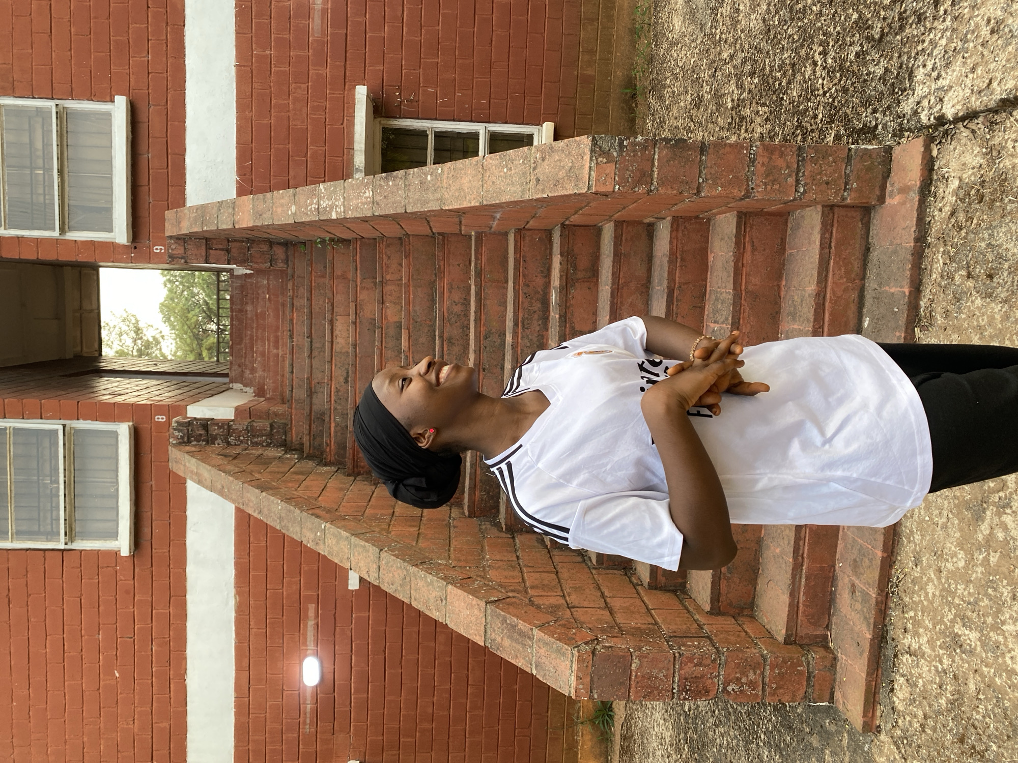 A young person, possibly female, stands outdoors, smiling broadly and looking upwards, with their hands clasped in front. They have dark skin and are wearing a white short-sleeved t-shirt with black stripes on the sleeves, indicative of an Adidas brand, and a "Fly Emirates" logo on the chest. They also wear a black head covering, dark pants, and a small red stud earring.
The setting is an outdoor area in Vom, Nigeria, featuring a red brick building with white trim. To the person's right, a set of brick stairs with matching brick railings ascends upwards. The building wall behind the person has a prominent vertical white stripe. Two windows with white frames and metal grates are visible on the left side of the image, set into the brick wall. The ground is a light, textured surface, possibly concrete. Through an opening beneath the stairs and building, trees can be seen in the background, suggesting lush outdoor surroundings. The lighting indicates it is daytime.
Notable details include small numbers "6" and "8" visible on the brick walls in different sections. The person's joyful expression and composed stance suggest they are posing for a photograph.
