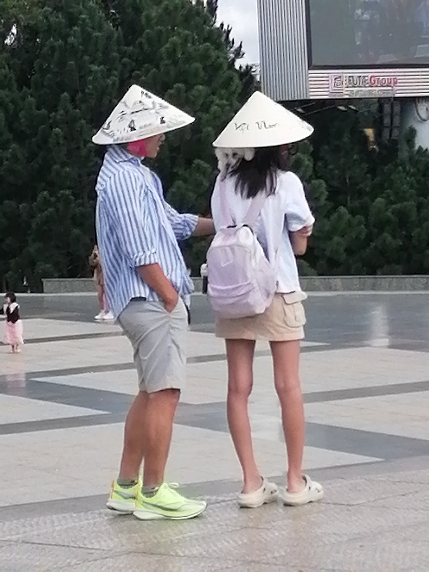 The image depicts two individuals wearing conical hats, a common sight in Vietnam. The person on the left is a male, wearing a blue and white striped shirt and grey shorts. He is standing with his hands in his pockets, facing the right. The person on the right is a female, wearing a white t-shirt and a tan skirt. She has a light purple backpack on her back and is facing away from the camera. The location is identified as Da Lat, Vietnam, an area known for its lush greenery and cooler climate. In the background, there are trees and a large screen, possibly a billboard or a display. The ground is paved with grey and white tiles, creating a geometric pattern. A small child in a pink dress is visible in the background to the left. The overall scene suggests an outdoor public space in Da Lat.
