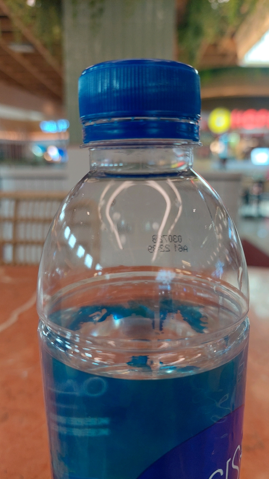 A clear plastic bottle with a blue cap is in focus. The bottle contains a blue liquid. Embossed text is visible on the side of the bottle. The background is blurred and shows a warm-toned interior with vertical wooden beams and shelves, suggesting a retail environment. The overall lighting is soft. The location is North Jakarta, Indonesia.