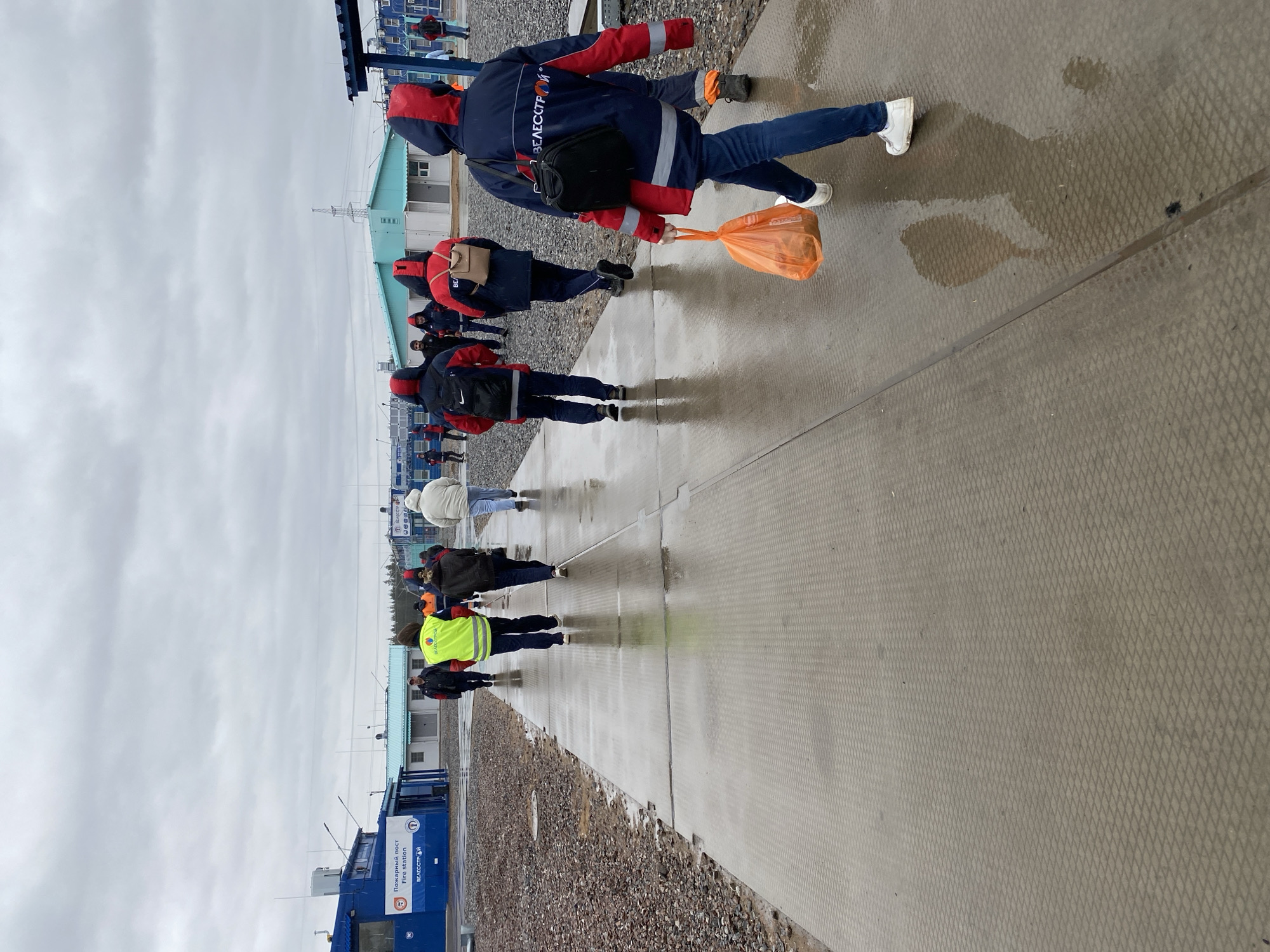 The scene shows a group of people walking on a concrete walkway next to a gravel area. The walkway appears wet, possibly from rain. Most of the individuals are wearing dark blue jackets with red hoods, some carrying bags. At the head of the group, one person is carrying an orange bag. In the background, there are blue and white buildings. A sign in Russian and English reads "Fire station". This indicates the location is Koskolovo, Russia.