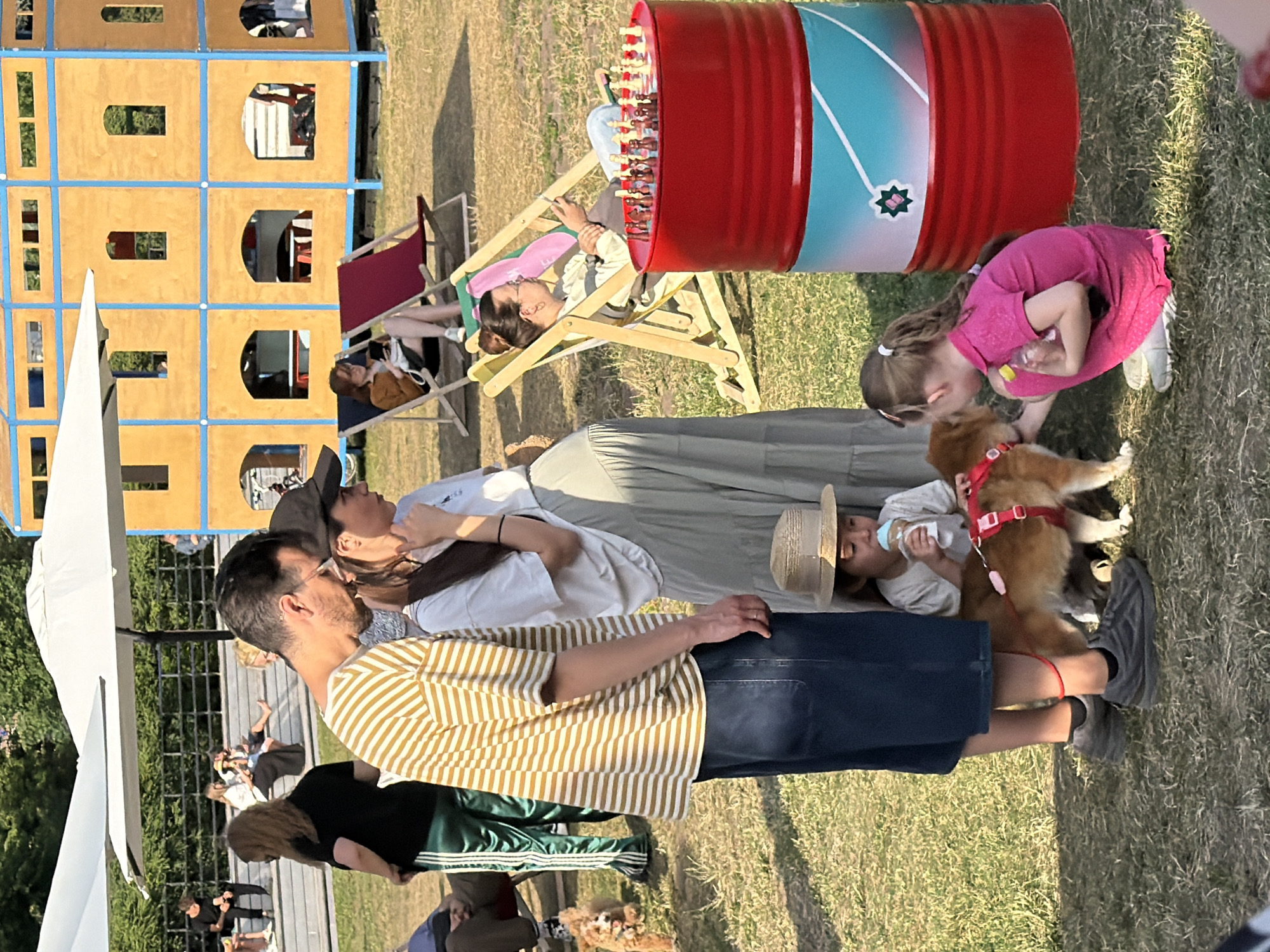 This image depicts an outdoor scene, likely a park or recreational area, with a group of people and a dog. In the foreground, a man in a striped t-shirt and dark shorts stands next to a pregnant woman wearing a white top and a long light-colored skirt. A young child with a straw hat is sitting down, holding a bottle, and interacting with a small, light brown dog. To the right, another young girl in a pink shirt is also interacting with the dog. Behind them, a large red barrel with a blue and white design is visible, and on top of it, a chessboard with pieces is set up. In the background, several other people are relaxing on lounge chairs, and a multi-story wooden structure resembling a playhouse or pavilion is present. The ground is covered in grass. The scene appears to be a casual gathering or event. There are no explicit visual cues in the image to indicate the specific city of Kazan, Russia.