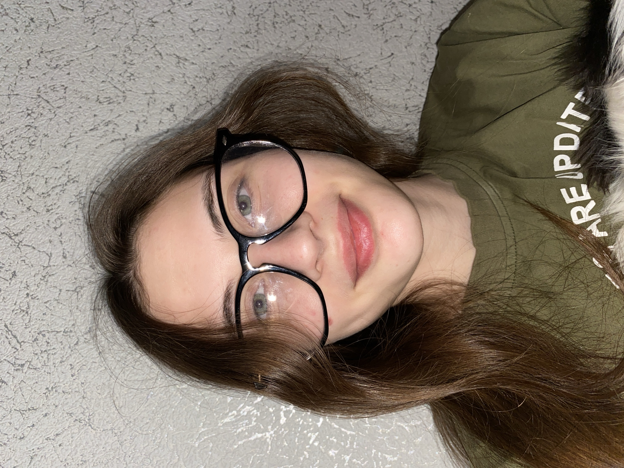 This close-up portrait, taken indoors in Votkinsk, Russia, features a young female subject. She is depicted from the chest up, seemingly lying down or leaning against a textured, light-colored surface, as the image is rotated counter-clockwise.
The subject has long, dark brown hair that frames her face and spreads around her head. She wears large, black-framed eyeglasses, with reflections visible on the lenses. Her eyes appear to be green or hazel, and she has a subtle, relaxed smile on her lips.
She is dressed in an olive-green long-sleeved shirt. Partial white text is visible on the right side of the shirt, which distinctly shows "ARE UPDIT", possibly part of a longer phrase like "ARE UPDATED". To the right of her head and shirt, a small section of white and black fur or fluffy material is visible, which could be from a pet or a clothing accessory like a hood.
The background consists of a uniformly textured, light grey or off-white surface, resembling a wall with a stucco finish or patterned wallpaper. The lighting appears bright and direct, creating reflections on her glasses and a general even illumination. The overall scene conveys a casual and relaxed atmosphere.