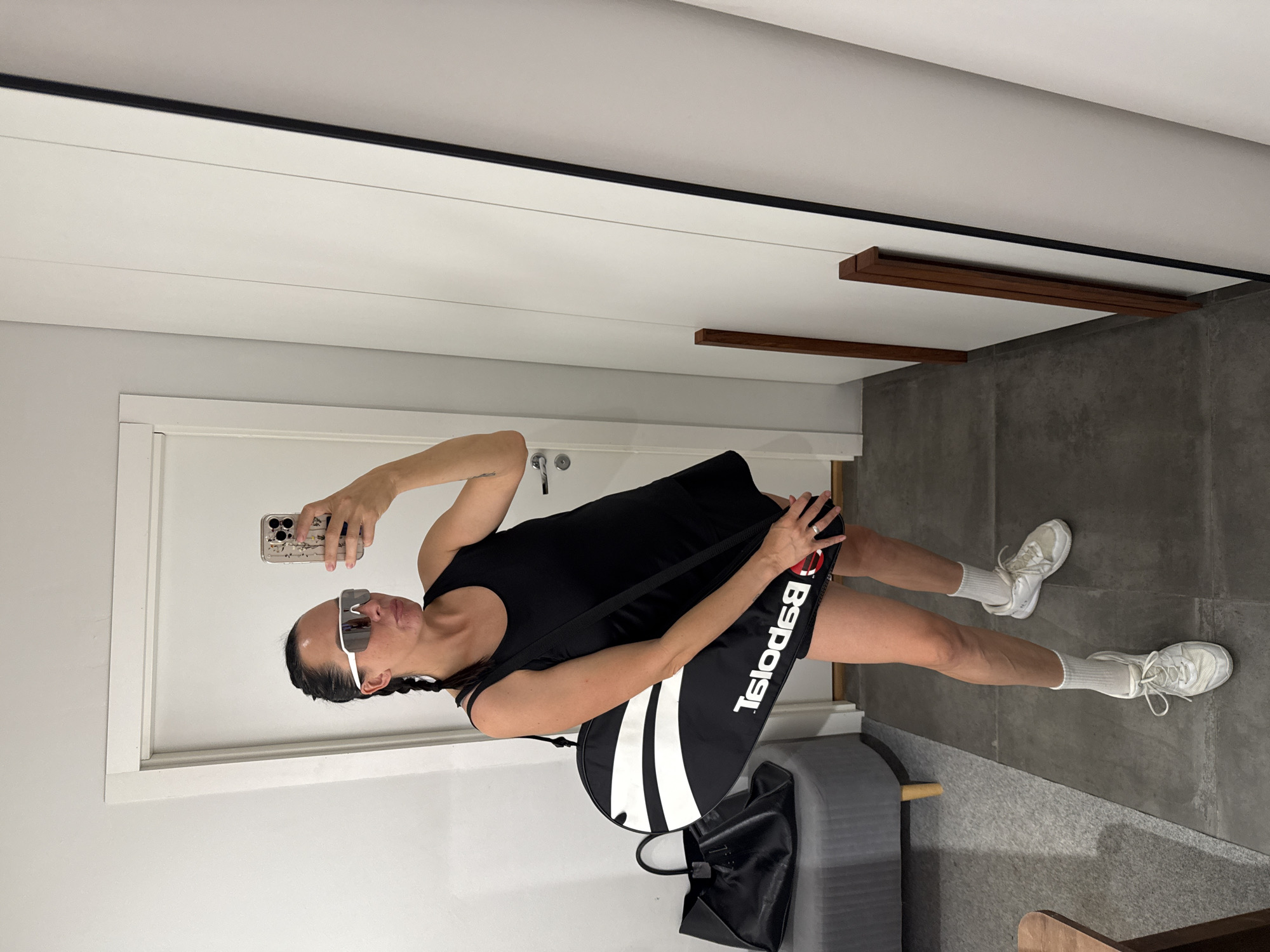 This media file shows a woman taking a mirror selfie. She is wearing a black tank top, black shorts, white socks, and white tennis shoes. She has her hair in two braids and is wearing large white sunglasses. She is holding a phone in her right hand, and in her left hand, she is holding the strap of a black tennis bag with white accents. The tennis bag has "BABOLAT" written on it in white, mirrored letters. Next to her is a black tote bag. The setting appears to be an indoor space with a white door and white walls. The floor is a dark gray tile. There are also wooden accents on the wall. The lighting suggests it is daytime, but there are no indications of the weather. The woman's expression is neutral.