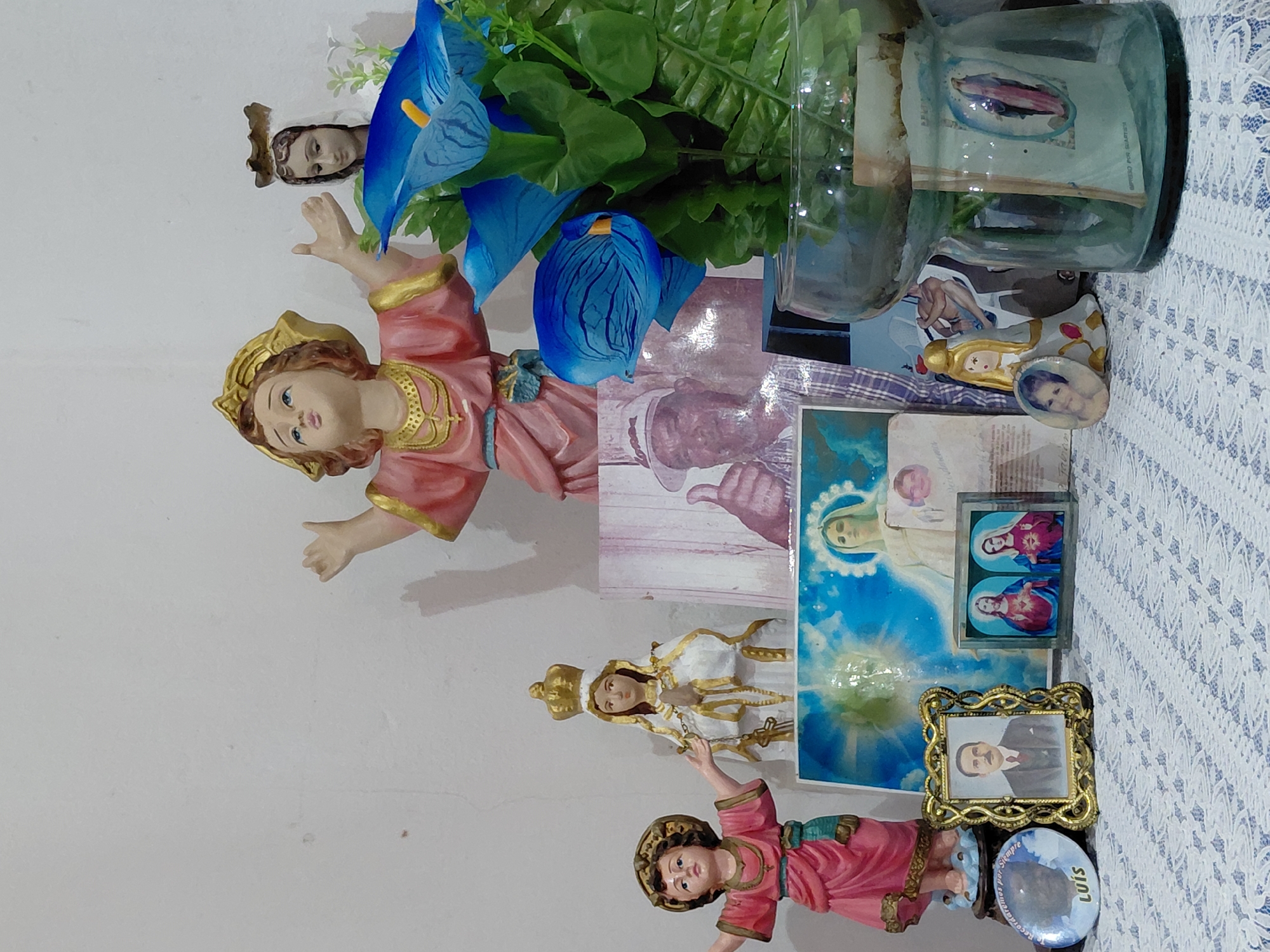 The image depicts an altar or shrine with several religious statues and photographs on a white, textured surface covered with a white and blue patterned cloth. In the center, a large statue of the infant Jesus in a pink and gold robe with outstretched arms is prominent. To its right, a statue of the Virgin Mary, also in white and gold, with a crown, is visible. Behind them, a large blue artificial calla lily arrangement with green leaves stands in a glass vase. Several framed photographs are also present, including a portrait of a man in a suit, a framed image of Jesus and Mary, and smaller circular photos. There is also a larger framed image that appears to be a religious depiction with a blue background. A glass jar containing greenery and a small religious image is on the right side of the frame. The background is a plain white wall. The setting appears to be an indoor domestic space.