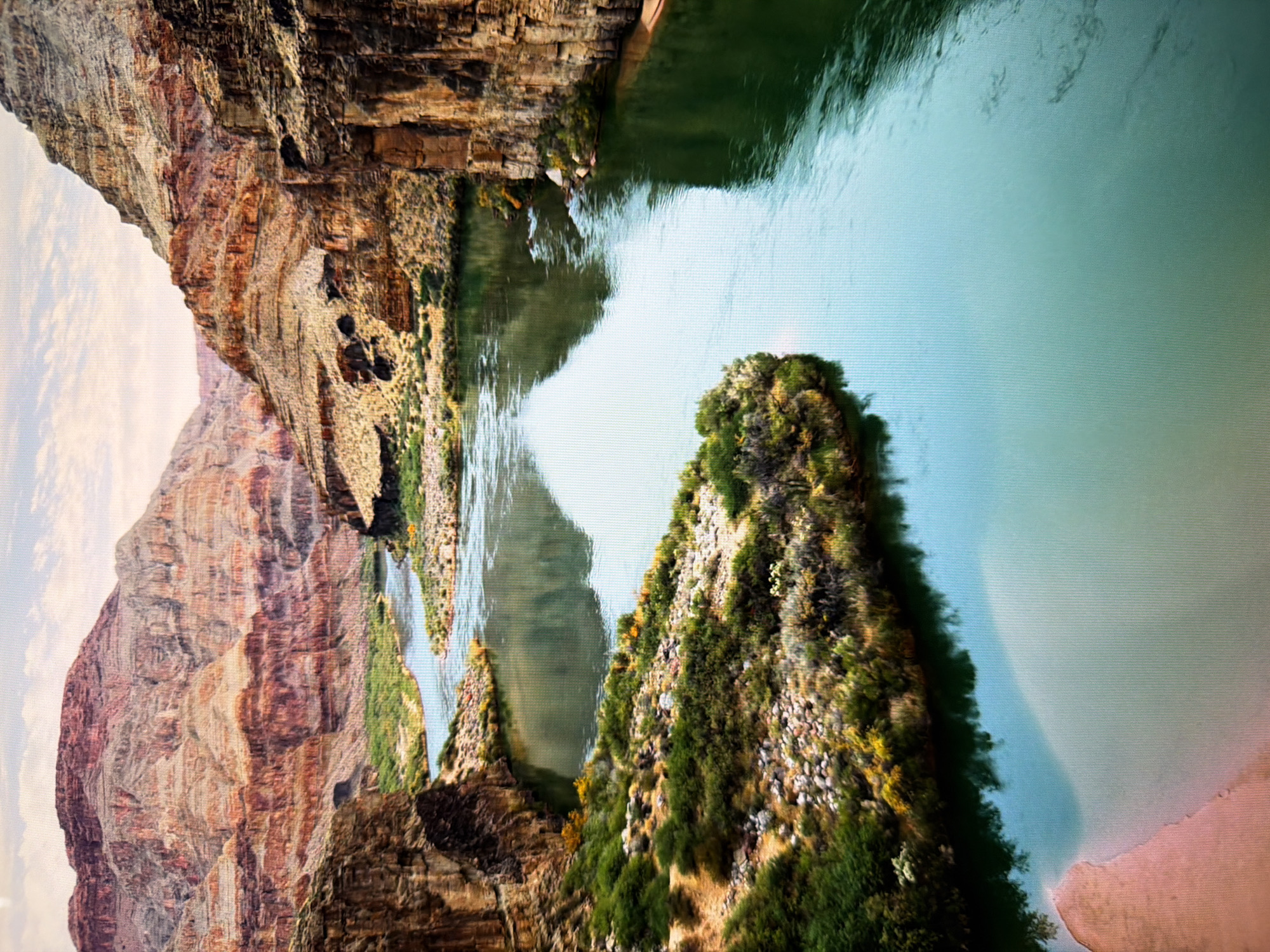 The image displays a wide, tranquil river flowing through a canyon. The canyon walls are steep and rocky, with layers of red and brown stone. Vegetation, including green bushes and trees, grows along the riverbanks and on some of the lower slopes. The water is a deep green color, reflecting the surrounding cliffs and the sky. In the foreground, a sandy bank with some pinkish hues is visible. The sky is partly cloudy with a soft, diffused light. The scene appears to be a natural landscape, likely a river canyon. There is no indication that this location is in Kyiv, Ukraine; it is more characteristic of a desert or semi-arid canyon region.