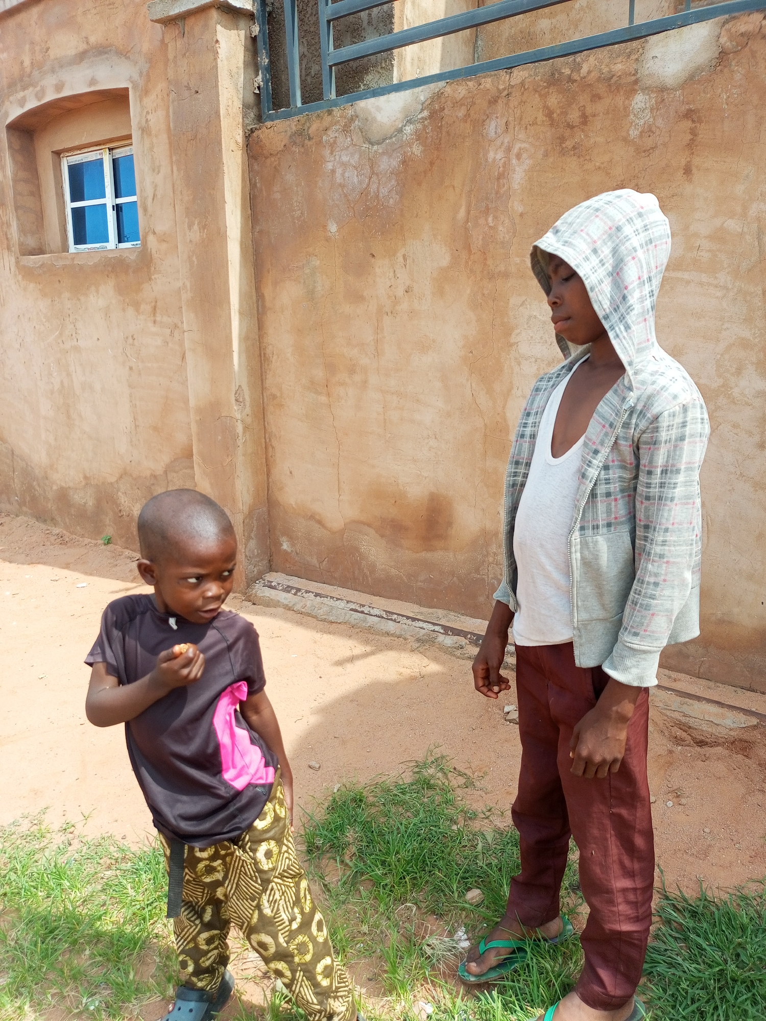 Two young boys stand in front of a weathered, ochre-colored wall in what appears to be a residential area of Gombe, Nigeria. The wall has a small, blue-paned window on the left and a section of dark metal bars at the top. The ground is a mix of reddish-brown soil and sparse green grass.
The boy on the left, younger and shorter, wears a black t-shirt with a bright pink design on the chest and patterned brown and yellow trousers. He is holding a piece of food up to his mouth and looking to his right. He has short hair and is wearing dark footwear.
The boy on the right, taller and wearing a hooded plaid jacket over a white tank top, stands facing the wall and looking down. His trousers are a reddish-brown color, and he is wearing green flip-flops. His hood is pulled up over his head. The lighting suggests it is daytime, possibly late morning or early afternoon, with shadows indicating the sun's position.