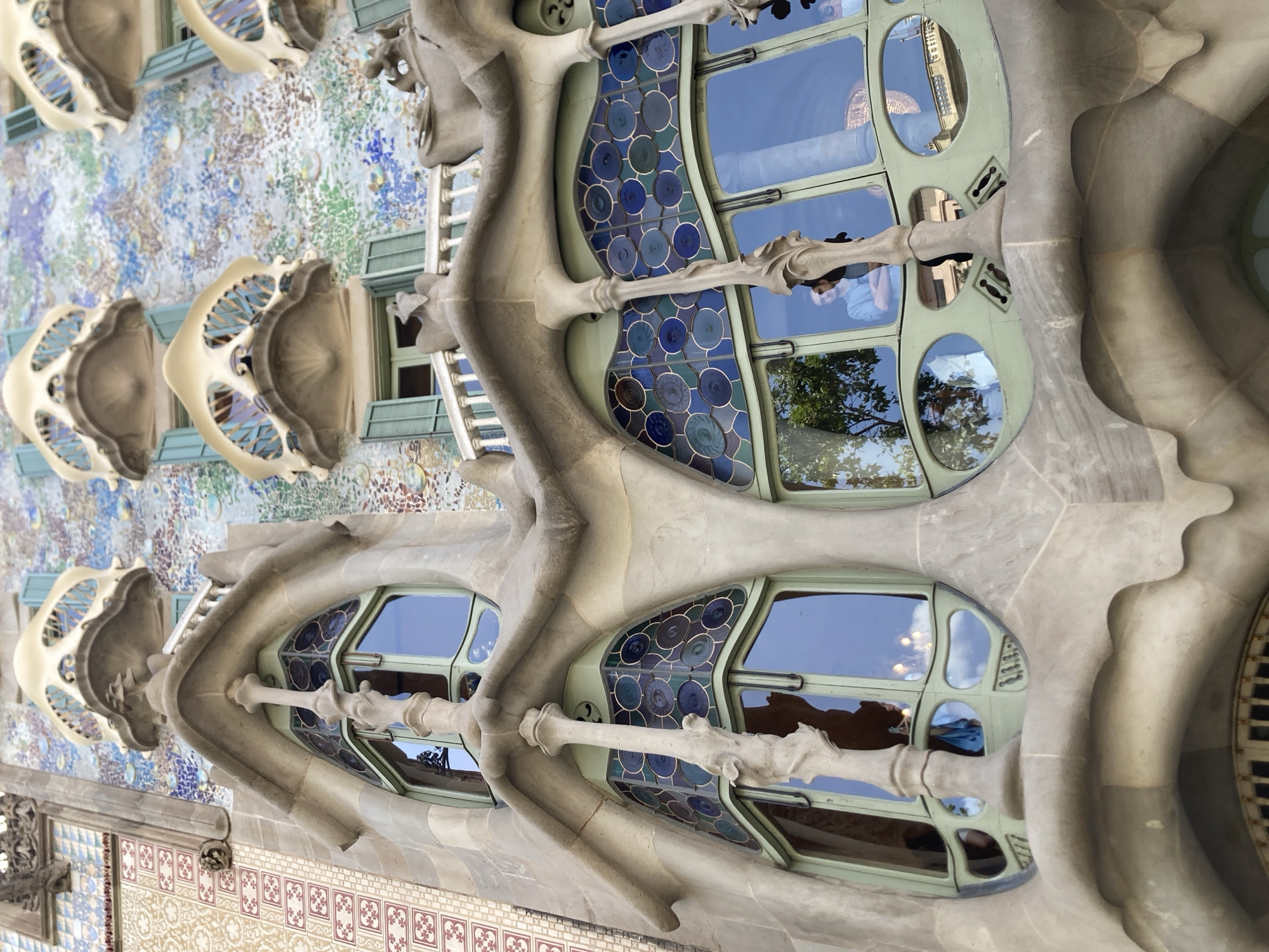 This is an exterior shot of a building with a unique, organic architectural style, likely Casa Batlló in Barcelona, Spain. The building's facade is covered in colorful mosaics and features flowing, bone-like structural elements. Large, curved windows with green frames dominate the scene, filled with stained glass in shades of blue and green that depict circular patterns. Reflections of the sky and surrounding trees are visible in the glass.
In the reflections of the windows, several people can be seen. In the upper window, a person in a light blue shirt is visible. In the lower windows, there are reflections of trees and the sky, as well as possibly other people and interior lights, suggesting it is daytime and possibly late afternoon or early evening. The overall impression is of a whimsical and artistic architectural design.