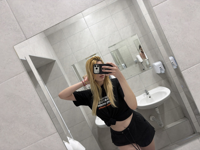 The image displays a blonde female taking a mirror selfie within a tiled restroom or changing room in Varna, Bulgaria. The subject, with fair skin and long straight blonde hair, is partially visible from the mid-thigh up. She is wearing a black t-shirt, knotted at the waist to expose her midriff and a navel piercing, and black shorts with a drawstring. Her right hand holds a black smartphone with a case, angled towards the mirror to capture the reflection. Her left arm is bent, with the hand positioned behind her head, and multiple colored bracelets are visible on her left wrist. Text on her t-shirt, partially obscured, reads "...OTION / NO... / VARNA," strongly suggesting the location is associated with "GYM MOTION VARNA," a fitness facility.
The environment, visible in the mirror's reflection, consists of walls clad in light gray or off-white rectangular tiles. A white ceramic sink with a chrome faucet is centrally positioned below a wall-mounted white liquid soap dispenser and a white hand dryer. A smaller, secondary mirror is visible in the background, reflecting a partially obscured figure, appearing as a male torso with light skin, possibly bare-chested. The primary mirror itself shows minor dust or smudges near its upper edges.
