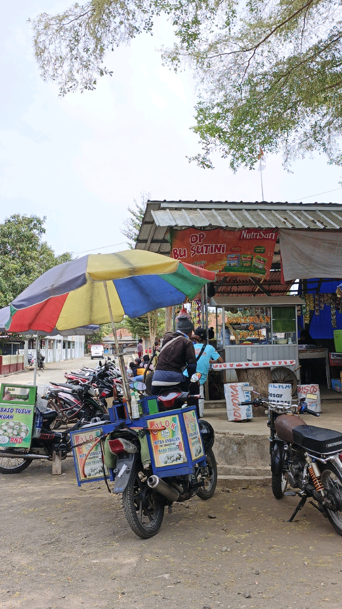 The image depicts an outdoor scene in Sukaresik, Indonesia, likely a roadside area near a food stall. In the foreground, a blue cart attached to a motorcycle is visible, advertising "BASO TUSUK KUAH". Another motorcycle with a similar cart, also advertising "BASO TUSUK", is parked to the left. A colorful umbrella, predominantly red, yellow, and blue, is set up to provide shade over the area.
Behind the umbrella, a food stall with a corrugated metal roof is operating. A banner above the stall reads "OP BU SUTINI" and advertises "NutriSari". The stall appears to be serving food, with customers visible in the background. One person, wearing a dark jacket and a beanie, is seated on a motorcycle in front of the stall. Another person in a bright blue shirt is standing at the stall. Several other individuals are seated or standing around the stall area.
In the middle ground, a row of parked motorcycles is lined up. Further back, a white vehicle is visible, and the environment suggests a rural or semi-urban setting with trees and vegetation. The ground is covered with dirt and gravel. The overall atmosphere is that of a busy street food vendor operation.