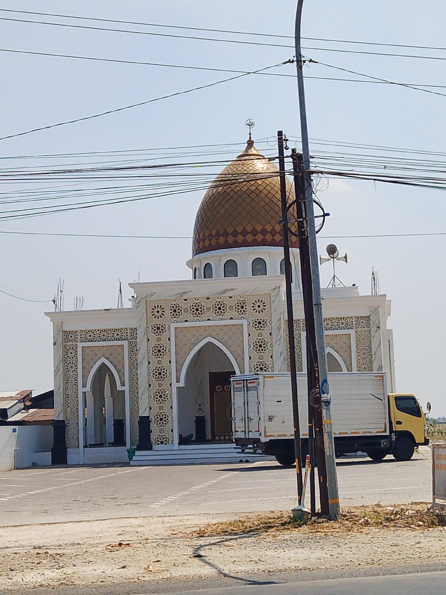 The image shows a mosque with a golden dome, located in the city of Bermi, Indonesia. A yellow truck is parked in front of the mosque. The sky is clear and blue, and there are several power lines visible in the foreground. The mosque has intricate geometric patterns on its facade and a crescent moon symbol on top of the dome. The truck is white with a yellow cab. The ground in front of the mosque is paved with asphalt and has white lines marking parking spaces. There is some vegetation and dry grass on the side of the road. The overall scene appears to be a sunny day in a relatively calm environment.