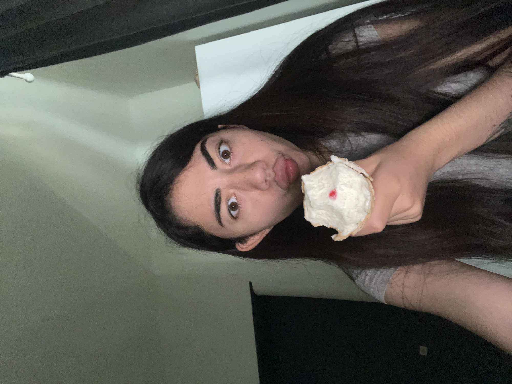 A young woman with long, dark hair is shown in a close-up, selfie-style shot. She is wearing a gray t-shirt and is holding a half-eaten ice cream cone with white cream and a red topping up to her mouth. Her lips are pursed, and her eyes are wide open, looking directly at the camera. The background is dimly lit, with a dark curtain on the right side of the frame and a white cabinet or wall on the left. The overall impression is of a casual, personal moment captured in a home environment. There is no indication of the image being located in Chotěšov, Czechia.