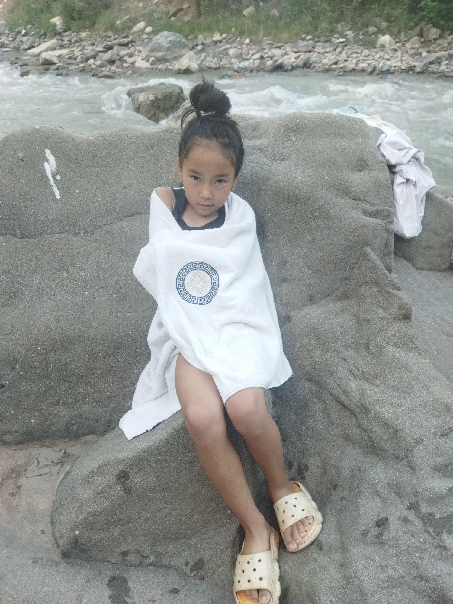 A young girl with her hair in a bun, wrapped in a white towel with a circular blue and white design, sits on a large gray rock by a river. She is wearing a dark colored tank top and light colored sandals. To her right, on the rock, are a few pieces of white clothing. The river is flowing with moderate speed, and the background shows a rocky shore with sparse vegetation. The lighting suggests it is daytime.