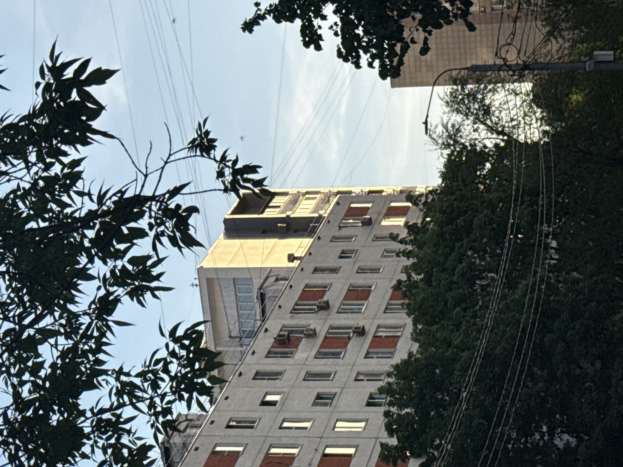 A low-angle view captures a tall, Soviet-era building in Moscow, Russia. The building is framed by tree foliage on both sides and the sky above. The building is rectangular with multiple floors, each featuring windows and red-colored window coverings. Some of the windows also have air conditioning units installed. A modern-style addition is visible on the top portion of the building, and there are power lines extending from the left side of the image. The sky is pale blue.