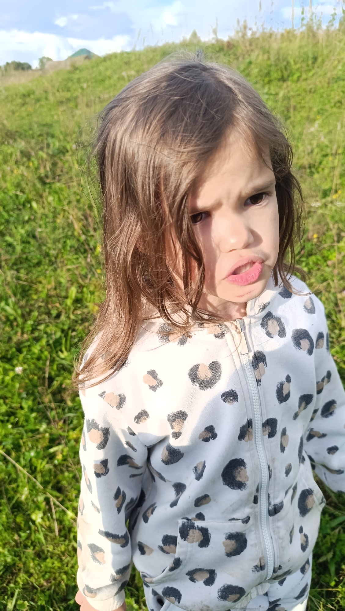 A medium shot captures a young girl with dark brown hair and a frustrated expression in an outdoor environment. She is wearing a white zippered hoodie with a leopard print pattern. The background consists of green grass and a slightly hilly landscape under a cloudy sky. There is no indication that this scene is located in Karsha, Russia.