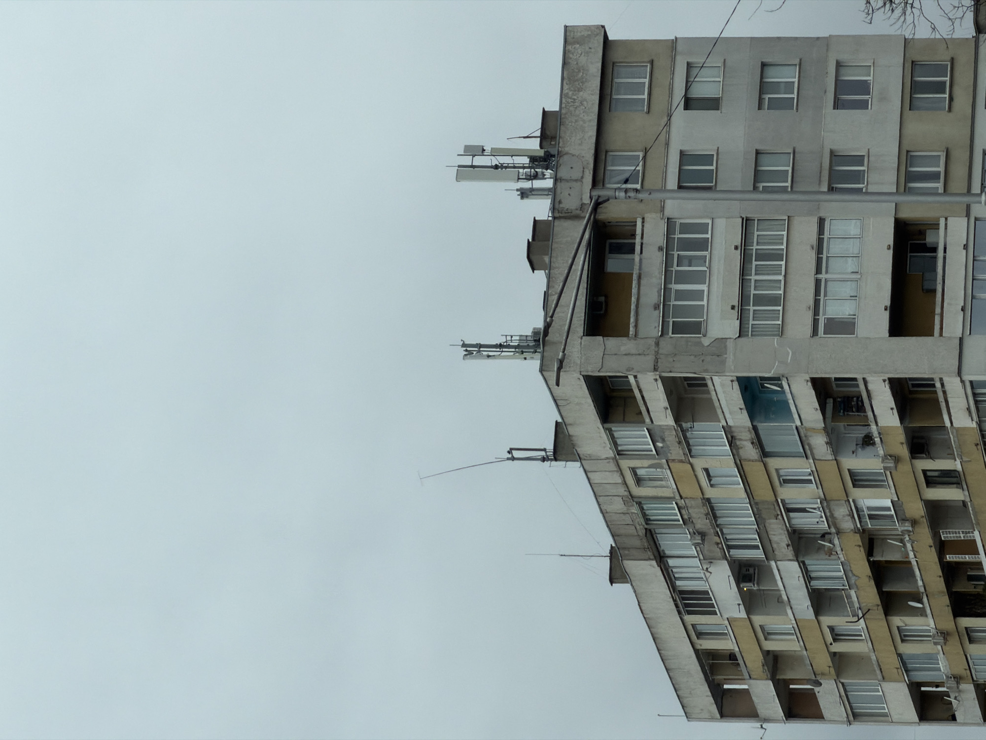 A tall, dilapidated concrete building, likely an apartment complex, dominates the right side of the frame. The building appears aged and poorly maintained, with visible cracks and discolored patches. It features numerous windows, some of which are open or missing, indicating disrepair. Various antennas and communication equipment are mounted on the roof and upper sections of the building. The sky is overcast and gray, filling most of the left side of the frame.
Given the style of architecture, it is plausible that this image depicts a building in Sofia, Bulgaria, as such structures are common in post-socialist countries. However, without further information, definitive location cannot be determined.