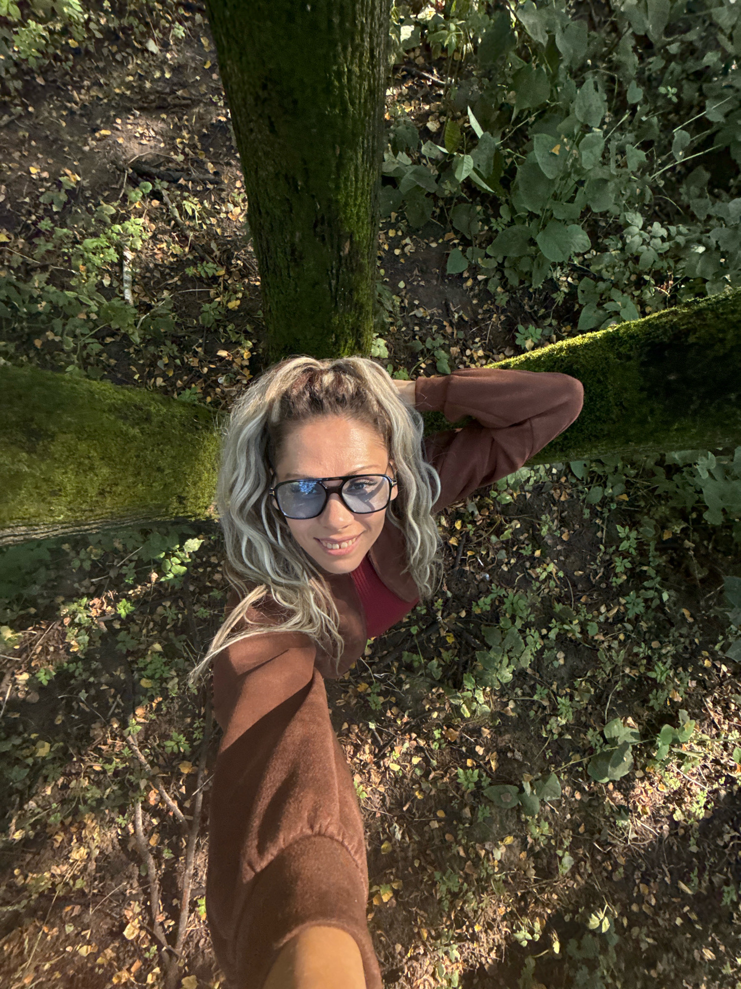 A woman is captured from a high angle, looking up and smiling at the camera. She has blonde, wavy hair pulled into a ponytail and is wearing blue-tinted sunglasses and a reddish-brown hooded jacket over a red top. Her left arm is raised, with her hand resting on the back of her head. She appears to be in an outdoor environment, likely a forest or wooded area, with several trees visible. The ground is covered in dirt, fallen leaves, and various green plants. Sunlight filters through the trees, creating patches of light and shadow on the ground. The location is identified as Rusanovo, Russia.