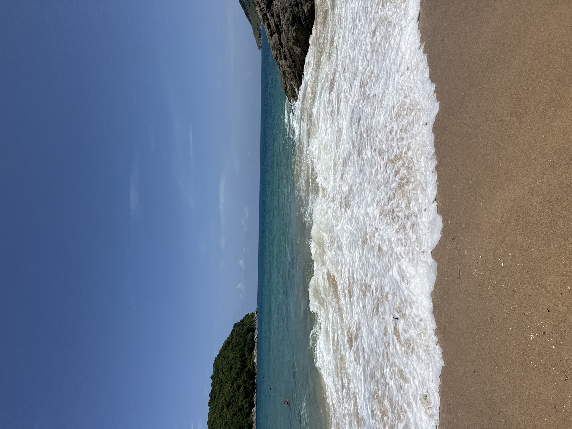The image depicts a serene beach scene in Rawai, Thailand, under a clear blue sky. In the foreground, gentle waves with white foam lap onto a sandy shore. To the left, a lush green island with dense vegetation juts out into the turquoise sea. Several people are visible in the water, some appearing to swim. On the right side of the frame, a large, dark rock formation marks the edge of the coastline. The overall impression is one of a peaceful tropical paradise.