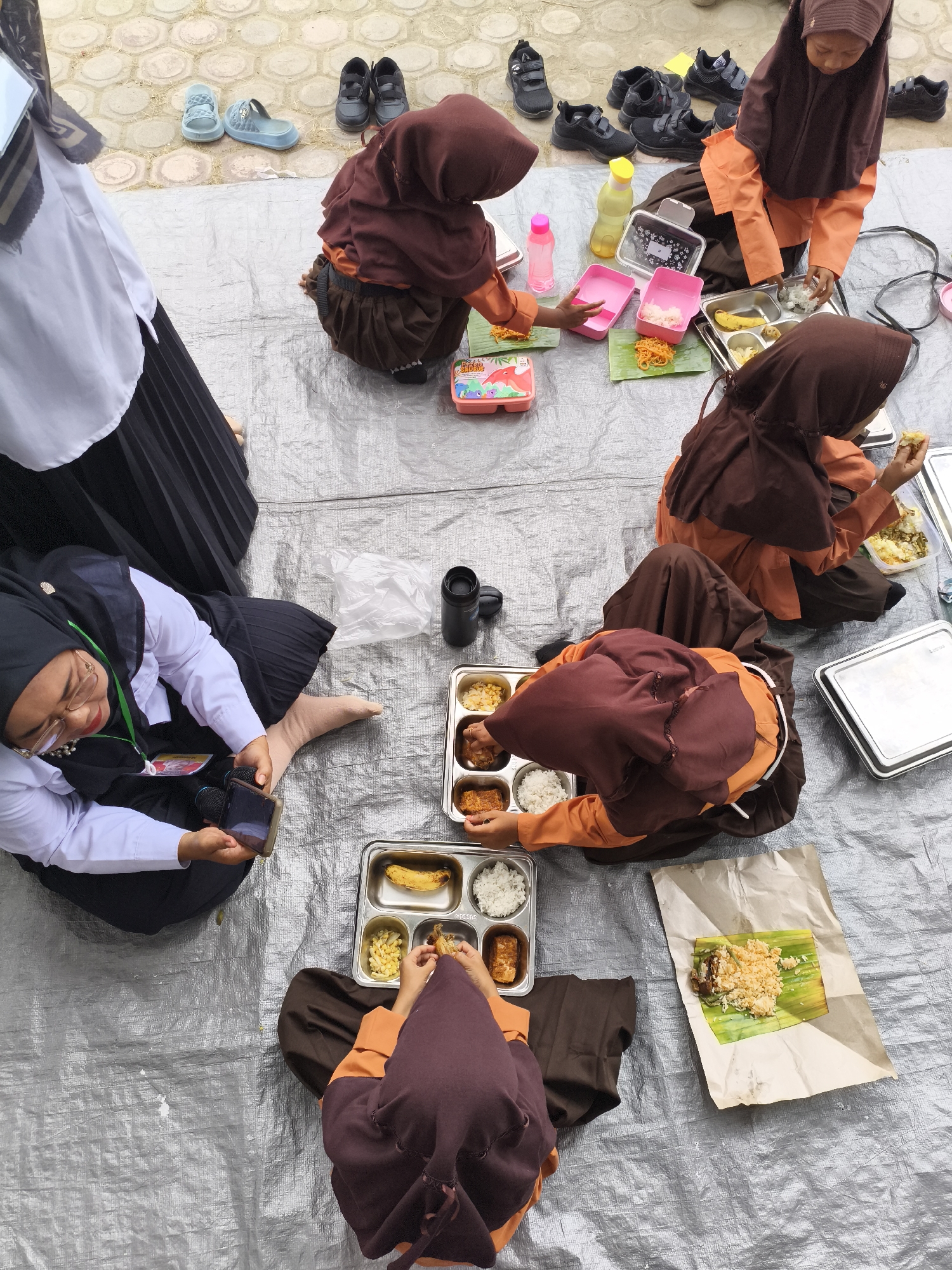 In Baratpayaitek, Indonesia, a group of young girls wearing modest Islamic clothing are seated on a grey tarp on the ground, engaged in eating. The children, primarily dressed in maroon hijabs and orange tops, are spread out with food containers and individual servings. Some are holding plates or trays, while others are directly eating from banana leaf-lined paper packaging. Among the scattered items are multiple pairs of black sneakers and a few blue sandals, suggesting this may be an outdoor school or community gathering. A woman in a white shirt and black skirt, also wearing a hijab, is seated on the tarp, looking at her phone. The background shows a textured, tiled ground. The scene appears to be a casual mealtime outdoors.