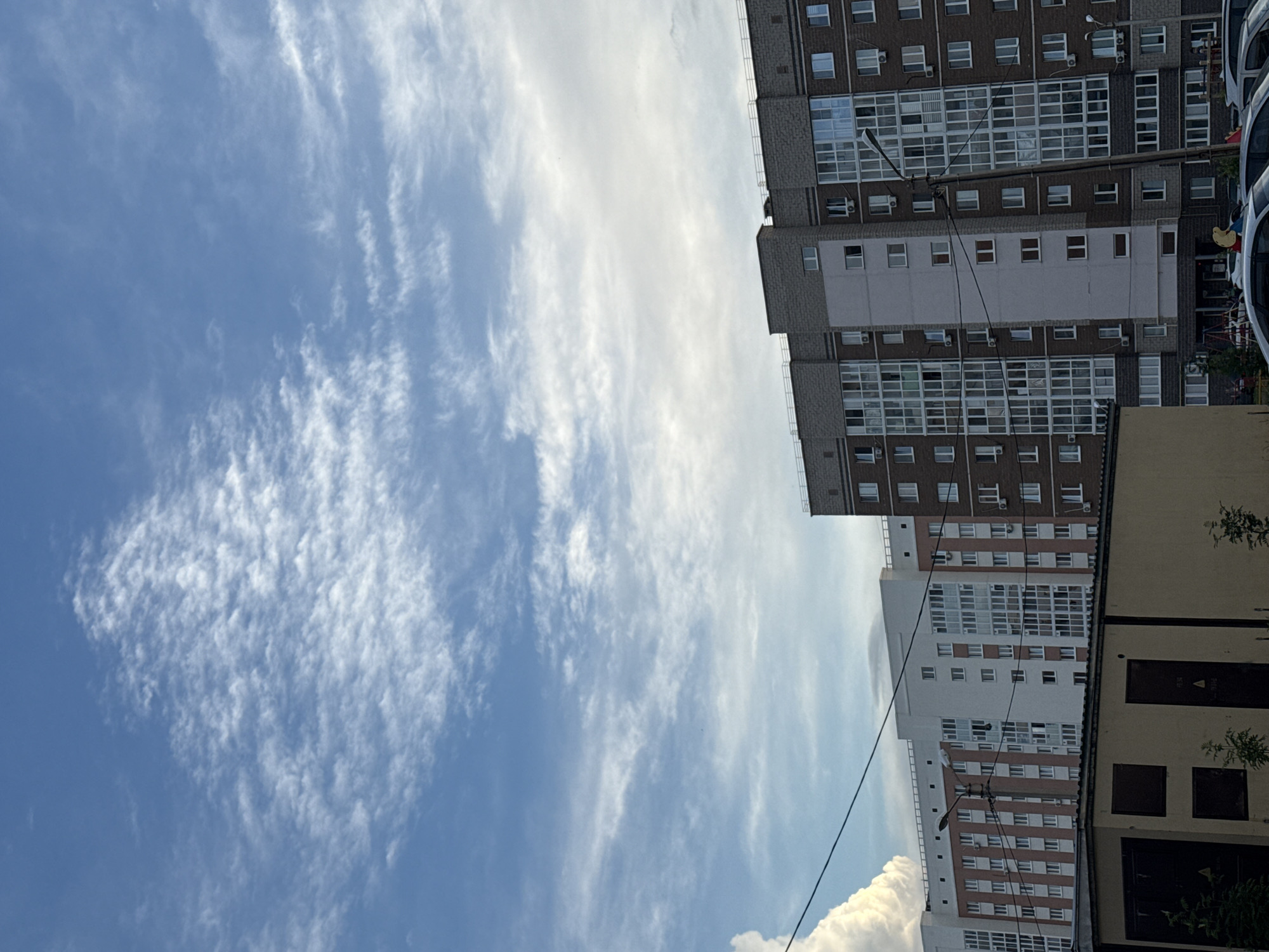 A medium shot captures a segment of an urban sky with a series of residential buildings in the foreground. The sky, a vibrant blue, is adorned with wispy white clouds that stretch across the frame, suggesting a bright day. Below the clouds, multiple apartment buildings dominate the lower portion of the image. The closest building is a tall, multi-story structure with a façade featuring a mix of white and pinkish-red brickwork, punctuated by numerous windows. To its right, another building showcases a darker brick exterior, also featuring a grid of windows and balconies. In front of these apartment blocks, a low-lying, tan-colored structure with dark garage doors occupies the very bottom of the frame, partially obscuring the bases of the taller buildings and the street level. The overall scene conveys a sense of urban living under a dynamic sky.
