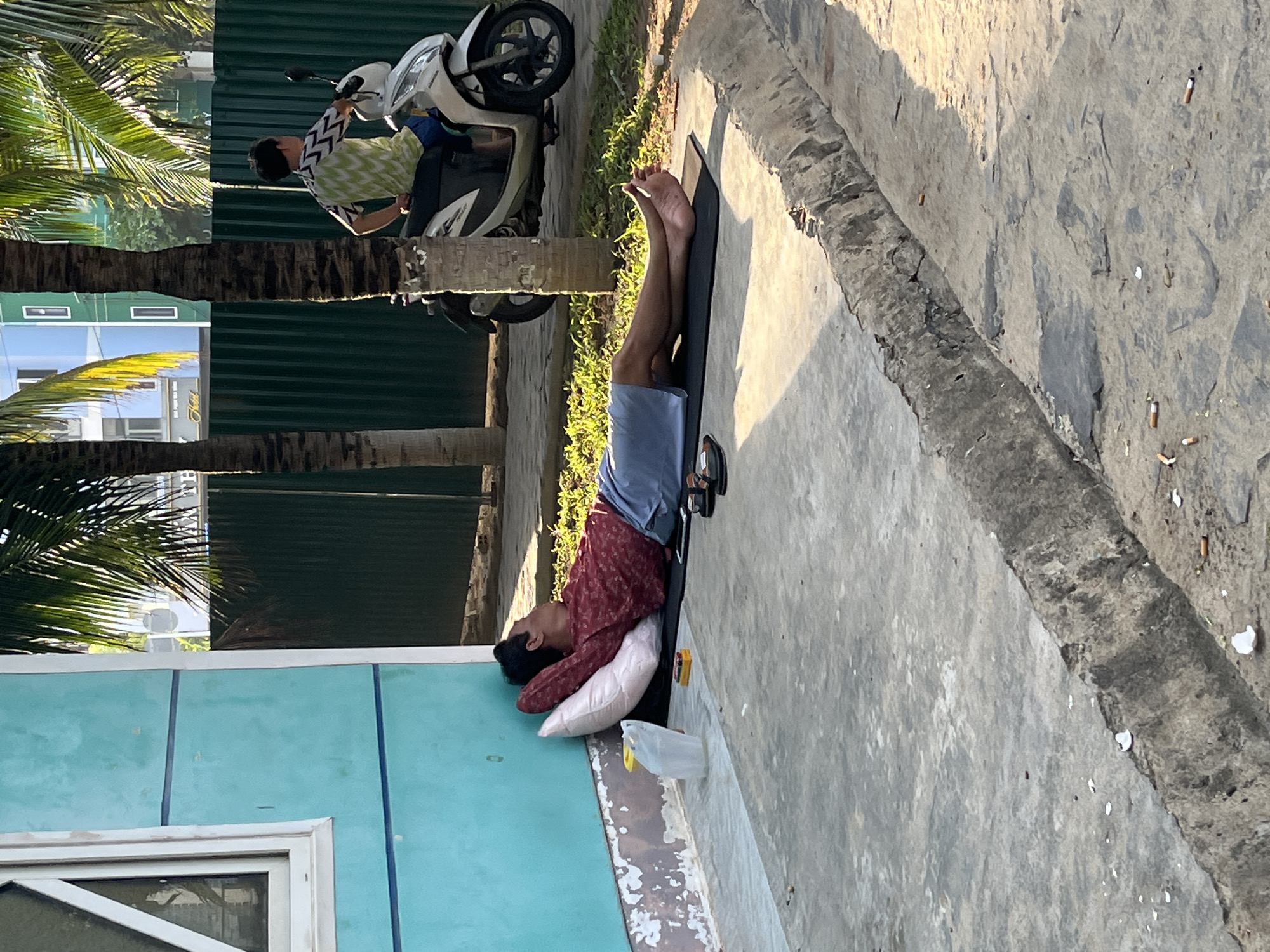 The scene captures a person lying on a black mat on a concrete surface adjacent to a building with turquoise walls in Bac Nha Trang Ward, Vietnam. The person is lying on their back with a white pillow under their head, wearing a red patterned shirt and blue shorts. A pair of sandals rests near their feet on the mat. Next to them, a white plastic bag sits on the concrete. The edge of the concrete surface has visible weathering and debris. Behind the building, a green corrugated metal fence runs alongside a row of palm trees. A white scooter is parked beside the fence. A person in a patterned shirt is crouched beside the scooter. Beyond the fence and palm trees, there is a glimpse of buildings. The overall impression is of an outdoor setting in the daytime.