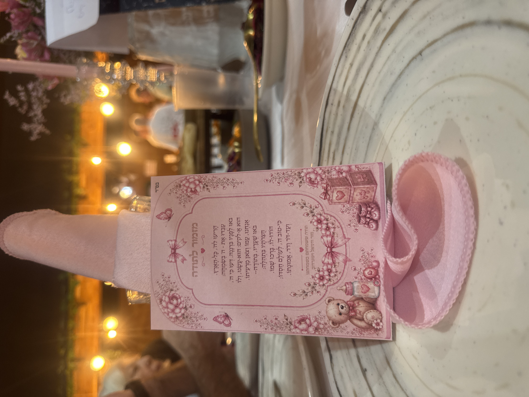 The image depicts a close-up view of a table setting with a pink decorative card featuring a teddy bear, baby bottle, and blocks. The card is placed on a white plate with a subtle patterned rim. A folded pink fabric is positioned on the plate in front of the card. In the background, blurred lights suggest an outdoor evening setting. While the image is centered on the table decor, it does not provide any visual information to indicate its specific location in Migdal HaEmek, Israel.