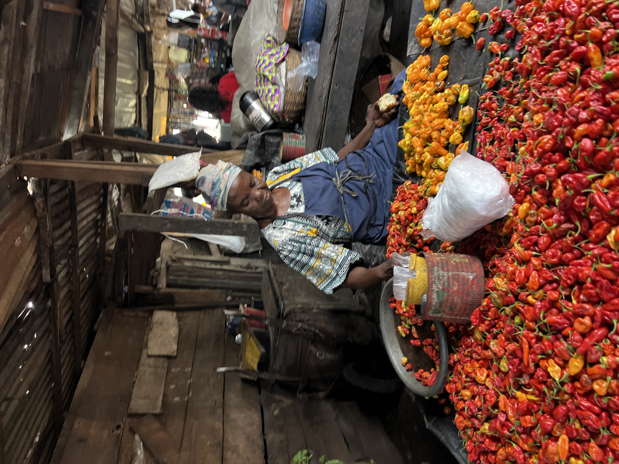 In Nkpor, Nigeria, a woman vendor is seen sitting at a market stall laden with a large quantity of red and orange peppers. She is wearing a patterned top and a blue apron, with a headscarf covering her hair. The stall is constructed from weathered wood, with a corrugated metal roof, typical of local market structures. Behind her, other vendors and shoppers can be seen in the background, suggesting a bustling market environment. A metal cart is visible to her left, and various containers, including a red and white tin and a plastic bag, are placed among the peppers. The overall scene depicts a vibrant market day in Nigeria, with a focus on the agricultural produce and the vendor's activity.