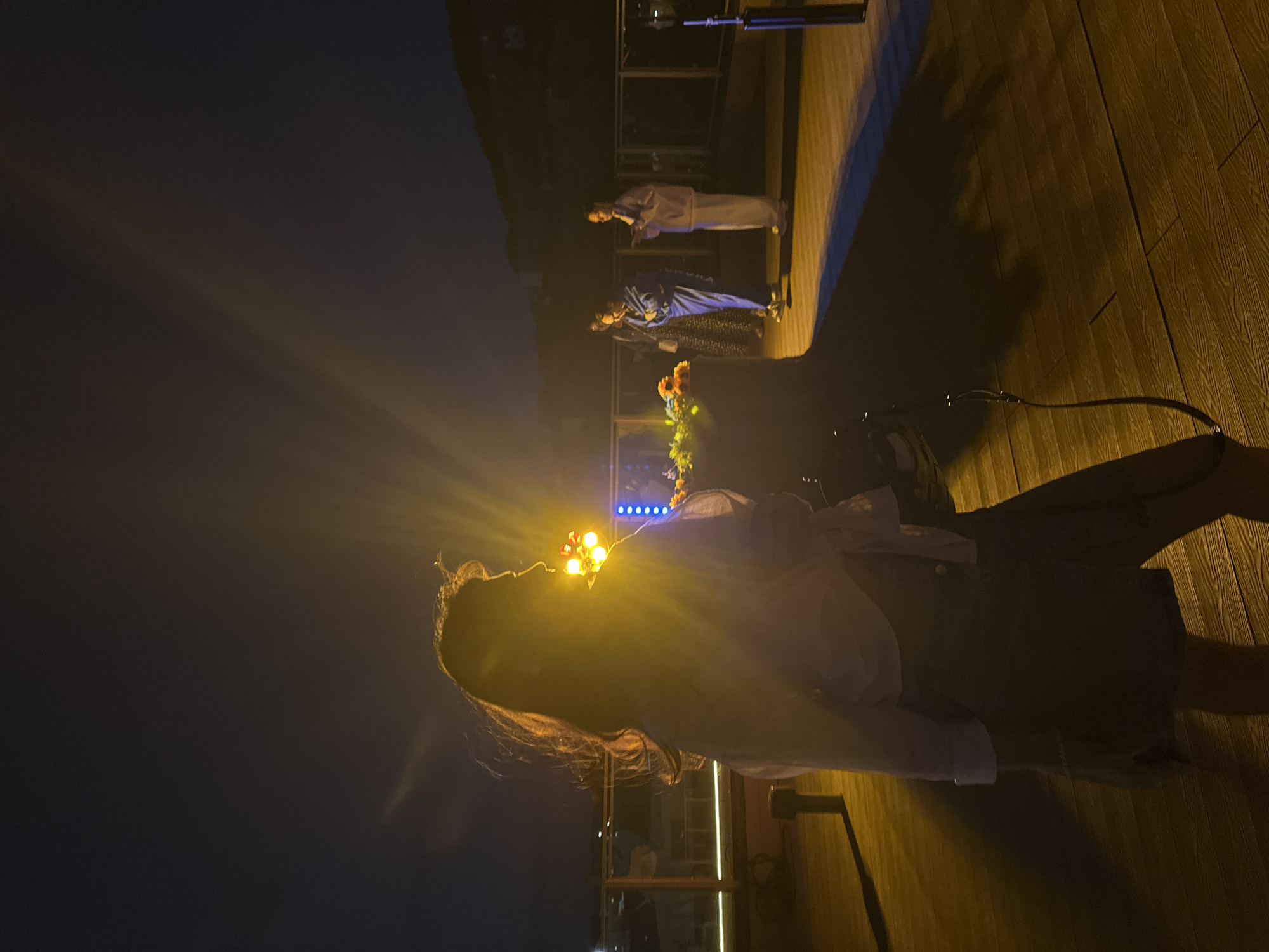 The image captures an outdoor nighttime scene, possibly a rooftop or balcony, in Petropavlovsk-Kamchatsky, Russia. In the foreground, a person is silhouetted against a bright, possibly artificial, light source. This person appears to be facing away from the camera, with their long hair illuminated by the light, casting a prominent shadow on the wooden deck.
In the midground, three individuals are visible. Two of them are wearing light-colored, possibly traditional or ceremonial, attire. The person on the left is wearing a blue garment and is looking down, while the person on the right is wearing a white outfit and holding what appears to be a piece of paper or a small book. Another person, in a patterned dress, stands behind them, slightly obscured.
A central feature in the midground is a large, dark object adorned with colorful flowers, primarily orange and yellow. Behind this floral arrangement, a string of blue lights is visible. The background shows a dark, hilly landscape, likely part of the city's natural topography, under a night sky. The overall ambiance is dimly lit, with the strong yellow light source in the foreground creating lens flare and accentuating the silhouette of the main subject. The wooden decking suggests an outdoor recreational or event space.