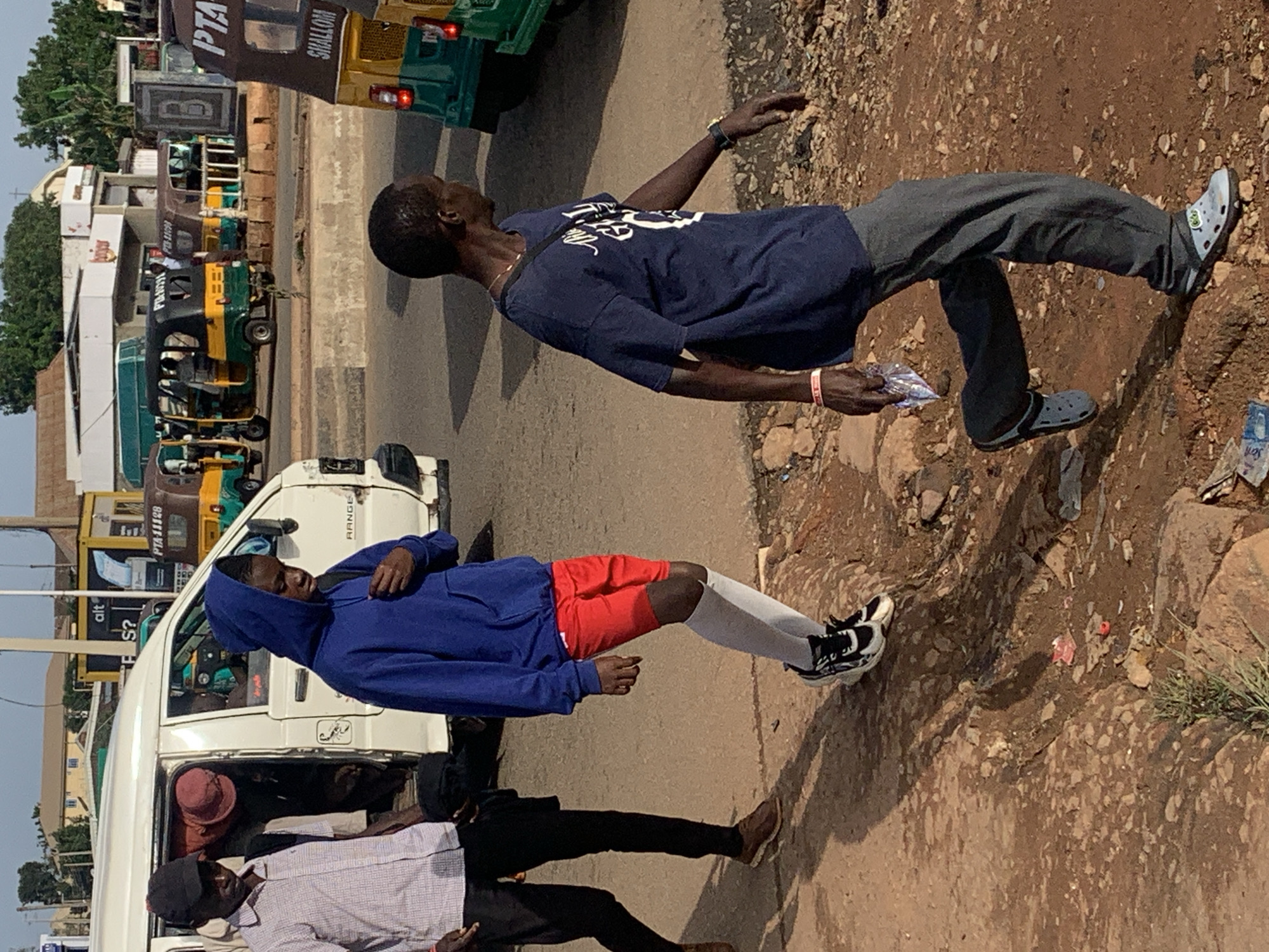 A group of people are standing on a dirt road next to a white van. The scene appears to be in Gura Top, Nigeria. One man, wearing a blue t-shirt and grey pants, is looking up and to his left, with his right arm extended. He is holding a small package in his hand. Another man, wearing a blue hooded sweatshirt and red shorts, is leaning against the van. He is wearing white socks and black and white sneakers. A third person, wearing a pink hat and a light-colored shirt, is visible inside the van. Another person in a black cap is standing next to the person with the pink hat. The ground is covered in dirt, rocks, and some debris. In the background, there are several auto-rickshaws, also known as "keke napep," and some buildings. The time of day appears to be daytime, and the weather seems to be sunny. Visible text includes "PTA 111G8" and "RANGE" on the white van, and "SHALOM" on one of the auto-rickshaws.
