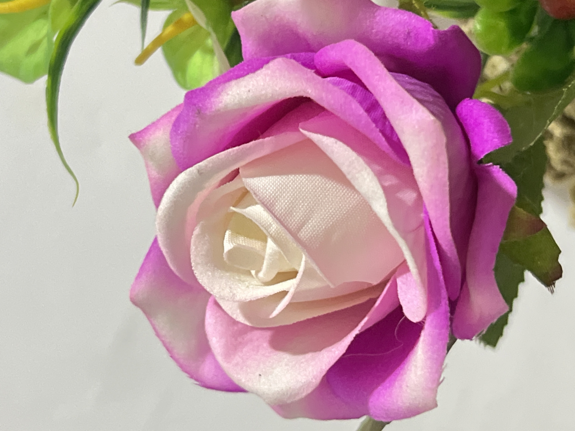 The image is a close-up photograph of a pink and white artificial rose. The rose is the primary subject, positioned centrally in the frame and taking up the majority of the image. The petals transition from a deep magenta on the outer edges to a lighter pink and then a creamy white towards the center. The texture of the petals appears to be fabric. In the upper right corner, out of focus, are some green leaves and a hint of yellow, suggesting it is part of a larger floral arrangement. The background is a plain, light-colored surface, likely a wall, which is slightly out of focus, further emphasizing the rose. There are no people, animals, or specific landmarks visible that would indicate a location within Denpasar, Indonesia. The lighting appears to be natural, coming from the upper left, casting subtle shadows on the petals.