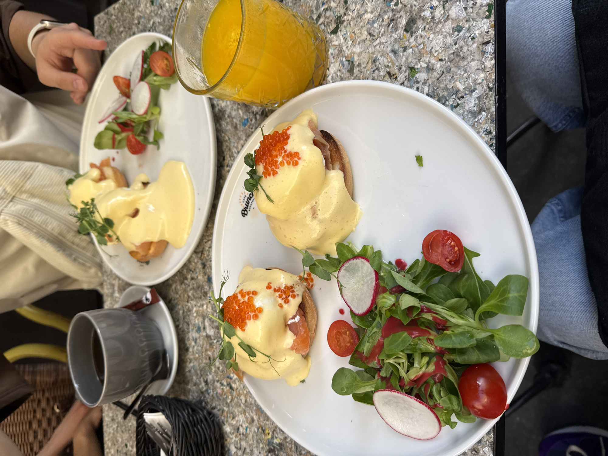 An overhead shot captures a vibrant brunch scene at a cafe, likely in Budapest, Hungary, with two plates of food, drinks, and partially visible patrons. The main focus is on a white oval plate featuring two servings of Eggs Benedict, each generously covered in creamy hollandaise sauce, topped with bright orange caviar, and garnished with delicate microgreens. The underlying protein beneath the sauce appears to be slices of smoked salmon. Accompanying the Eggs Benedict is a fresh side salad composed of mixed greens, slices of red radish, and cherry tomatoes, all dressed with a reddish vinaigrette. A second, partially visible plate in the background shows a similar presentation.
Drinks on the table include a large clear glass filled with bright orange juice and a grey ceramic mug with a dark beverage, likely coffee or tea, resting on a matching saucer with a spoon. Parts of two individuals are visible: one hand with a white bracelet reaching towards a plate, and legs, one clad in blue jeans, under the table next to a dark-cased mobile phone. The table surface has a unique, speckled texture, appearing to be made from composite or recycled materials. Cutlery is neatly arranged in a dark woven basket. Visible text on the rim of the main plate reads "Cafe Bruno BRUNCH," identifying the establishment. The bright, natural lighting suggests the meal is taking place during the daytime.