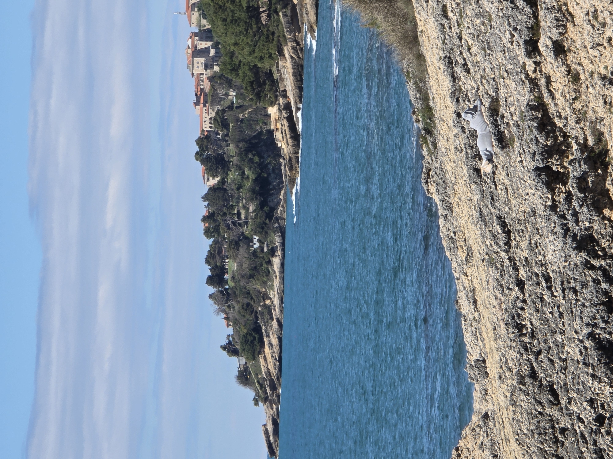 The image showcases a rugged coastline alongside the city of Ulcinj - Ulqin, Montenegro. A sheer rock cliff dominates the foreground, exhibiting a rough texture. Adjacent to the cliff is the turquoise sea. Across the water, the city of Ulcinj - Ulqin is nestled amid trees. The sky is visible as a bright blue expanse with elongated clouds. A person, dressed in light-colored clothing, appears to be climbing the cliff face.