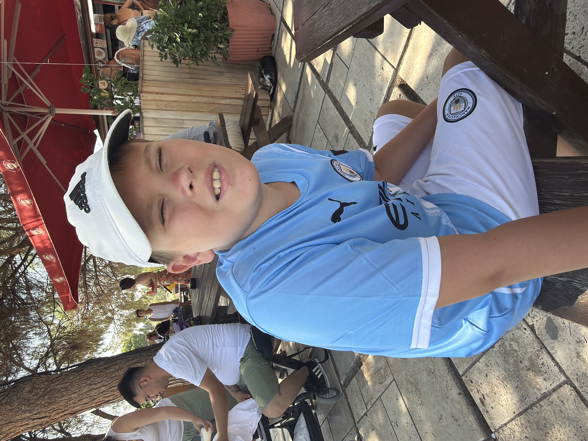 The media file shows a group of people outdoors, possibly at a cafe or restaurant with an umbrella providing shade. The primary subject is a young boy in the foreground, wearing a light blue jersey with a crest on it and white shorts with a similar crest. He has a white baseball cap on and is looking up with his eyes closed and a slight smile. The setting appears to be a paved outdoor area with wooden tables and chairs. In the background, other people are visible, some seated and some standing, enjoying their time. There is greenery and trees surrounding the area, suggesting a warm, sunny day. A cat is also seen lying on the ground near a potted plant. The crest on the boy's jersey and shorts appears to be the Manchester City Football Club logo. The overall mood seems relaxed and casual. The time of day is likely daytime, given the sunlight and shadows. The location is identified as Pamukkale, Turkey, which is known for its travertine terraces and thermal waters, though these features are not directly visible in this particular frame.