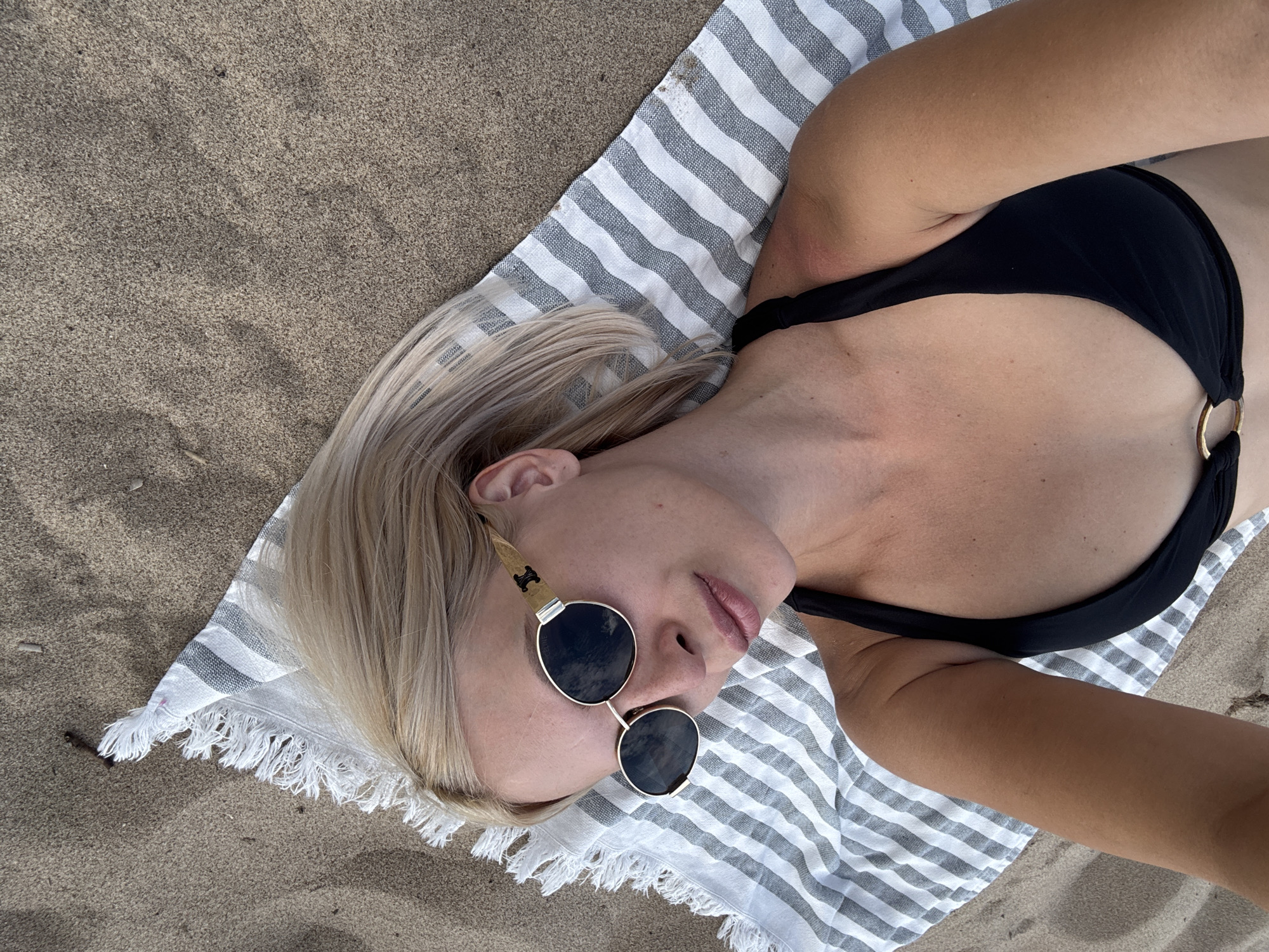 This media file features a person lying on a striped towel on a sandy beach. The subject is wearing a black bikini and sunglasses. The setting appears to be a beach, with sand clearly visible. The time of day seems to be daytime, likely sunny given the person is at the beach. The person's pose suggests relaxation and enjoyment of the environment. No visible text is present in the image. The image is taken from a high-angle perspective, looking down at the subject.