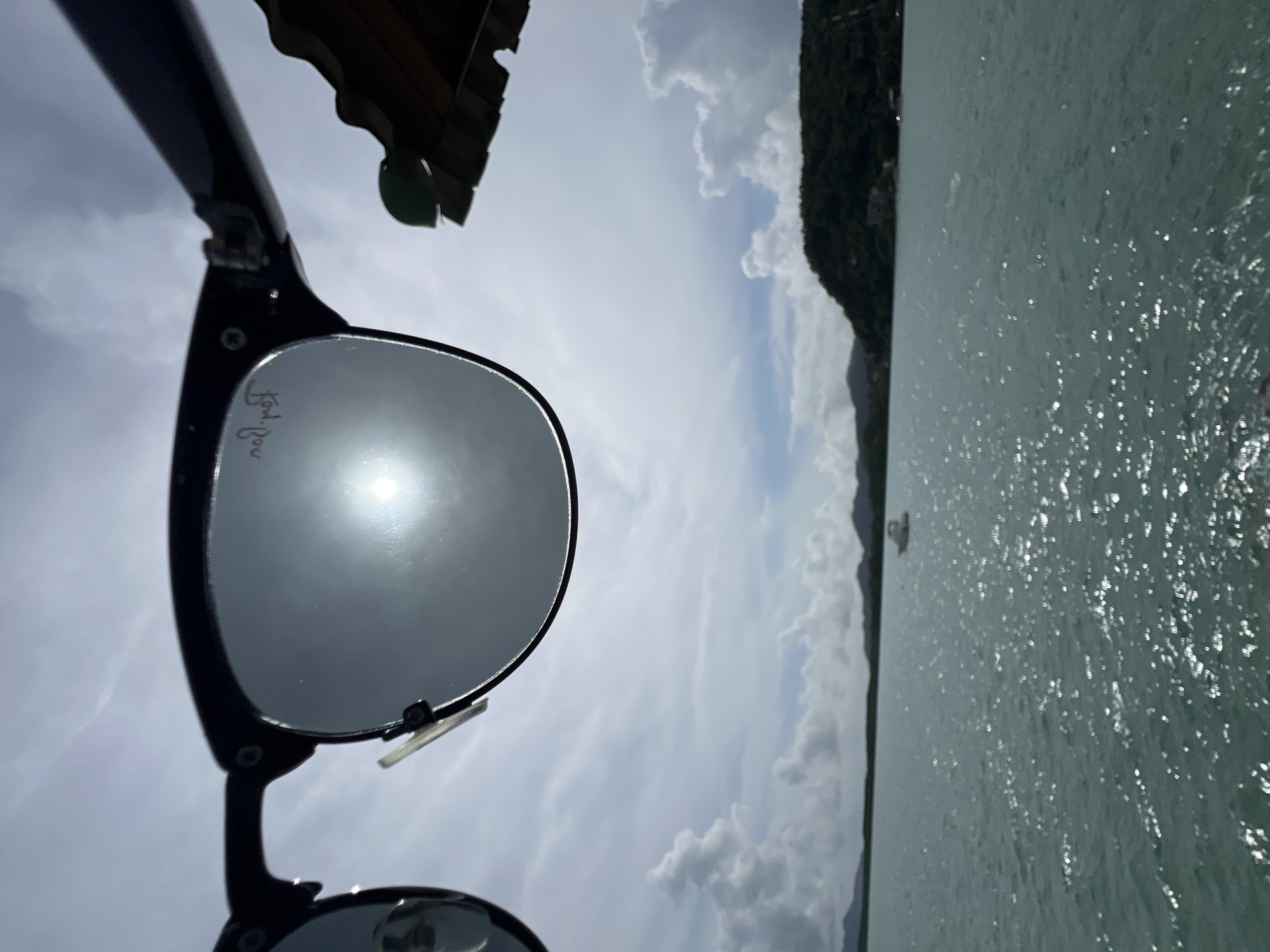 The image shows a close-up of sunglasses with the sun and sky reflected in the lenses. Beyond the sunglasses, a body of water is visible with a small boat in the distance. A lush green hillside forms the background, and fluffy white clouds dot the blue sky. The scene appears to be located in Pa Khlok, Thailand, a coastal area known for its natural beauty and calm waters.