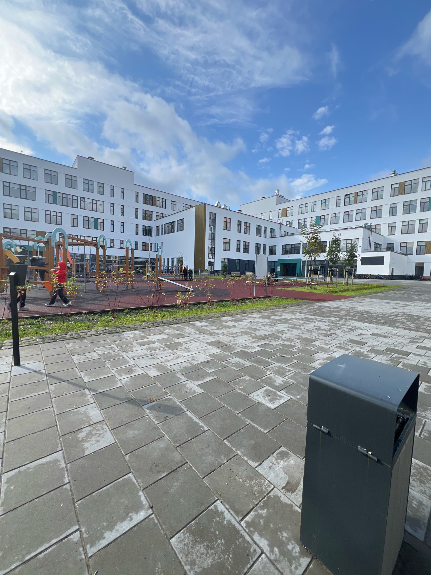 The image shows the exterior of a modern school building in Yekaterinburg, Russia. The building is multi-story and features a white facade with numerous windows. In the foreground, there is a paved courtyard with gray square tiles, some of which appear wet. A dark gray trash receptacle is visible on the right side of the courtyard. Beyond the courtyard, there is a children's playground area with various structures, including swings and climbing frames, on a red rubberized surface. Several children and adults are present on the playground, engaged in activities. Green grass and small trees are scattered throughout the scene. The sky above is blue with scattered white clouds.